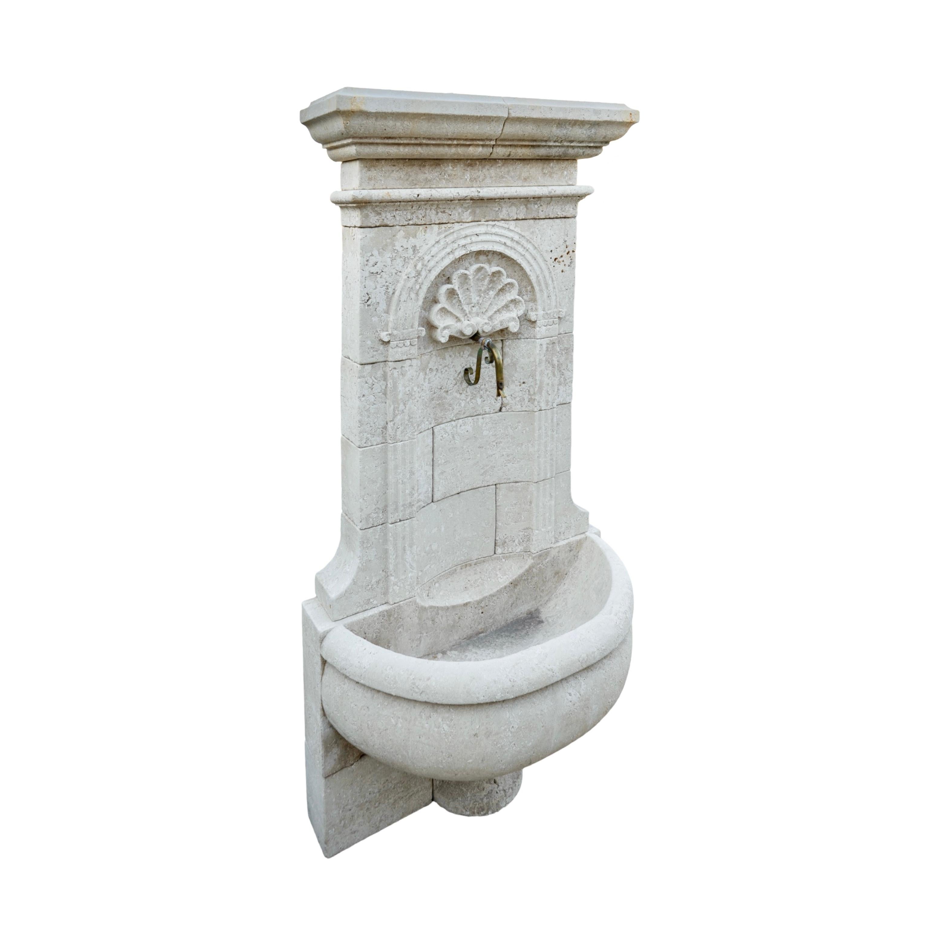 Une fontaine murale en pierre calcaire sculptée à la main en France, fabriquée dans un style contemporain. Cette fontaine comporte deux bassins : un petit bassin supérieur qui recueille l'eau du bec situé au-dessus et qui se déverse gracieusement