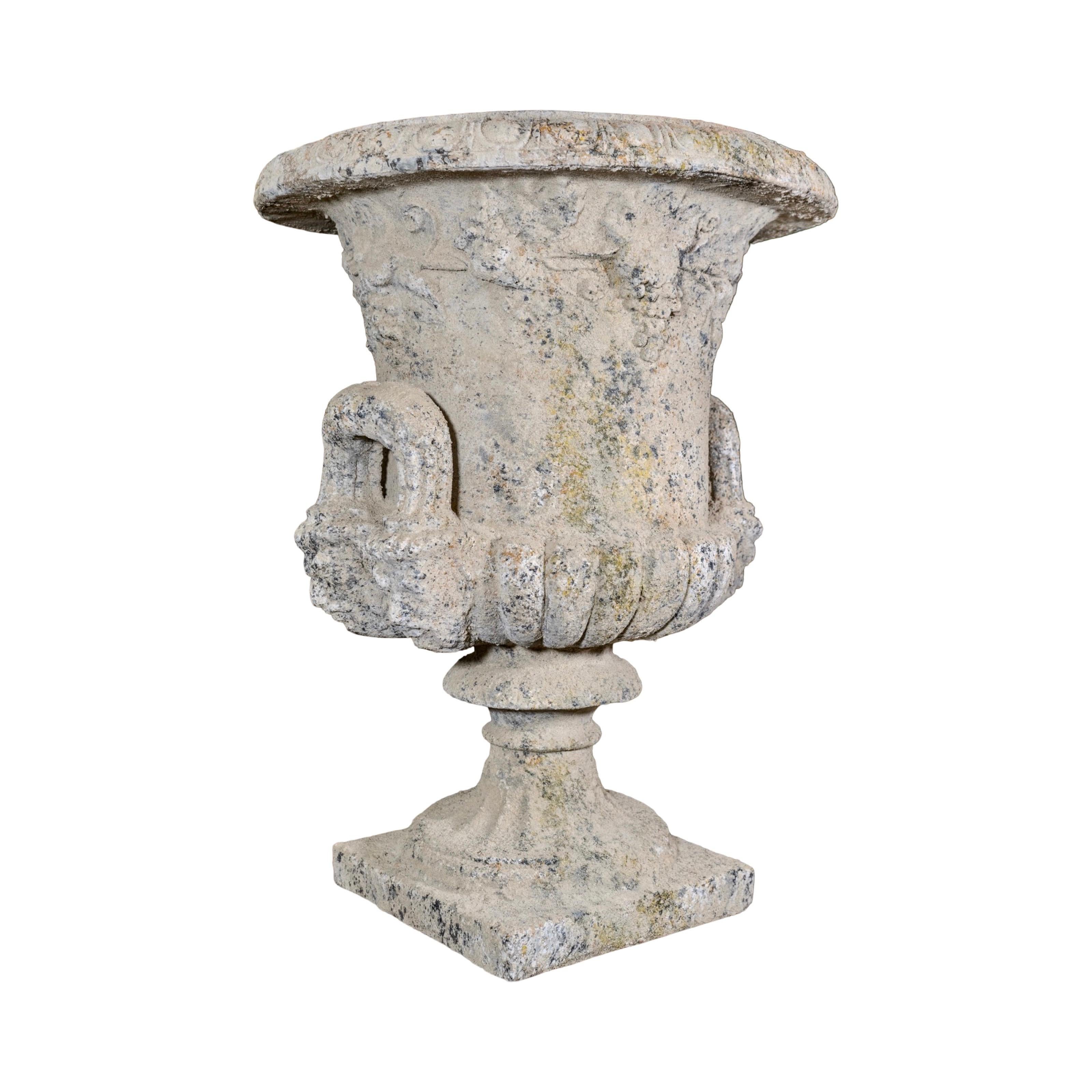 Un grand vase de style Médicis, richement orné de vignes et de raisins sculptés entourant son corps. Le bord décoratif présente des détails complexes, tandis que deux poignées latérales sont couronnées de visages de Bacchus sculptés à leur base.