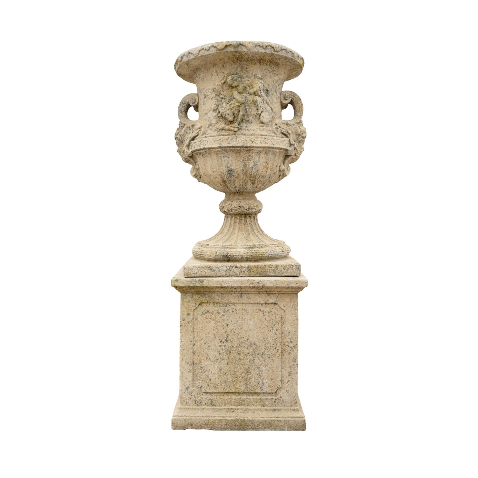 A contemporary French urn and base set made from reconstituted stone with an aged finish. The urn features scrolling handles and carved relief detailing, paired with a paneled base for a classic look. Large in scale, it can be planted with florals