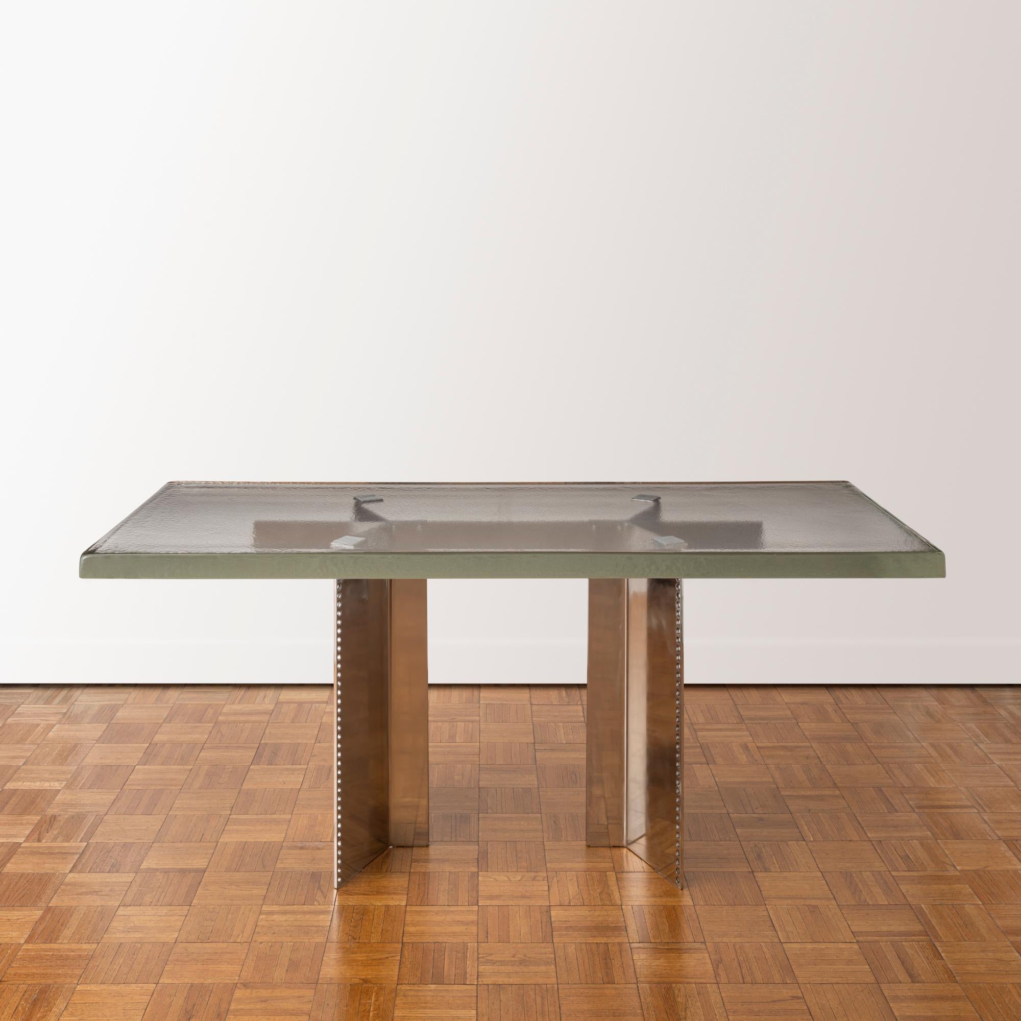 Contemporary dining table comprised of a rectangular glass slab atop an architectural base of perforated stainless steel pedestals joined by a stainless steel crossbar. The massive, two-inch thick glass top is produced by crucible casting, which