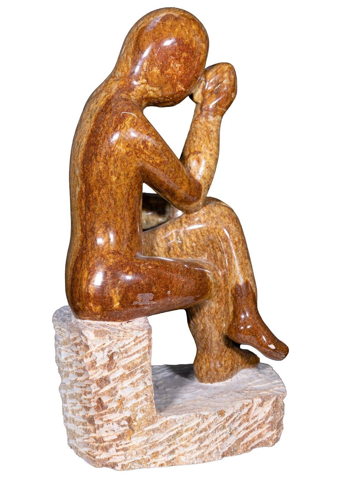 A glass soapstone figurine. This gorgeous sculpture depicts a figure sitting while in deep contemplation. This piece is made from a stunning polished brown soapstone and sits on a separate stone base. The gradations in the soapstone provide for a