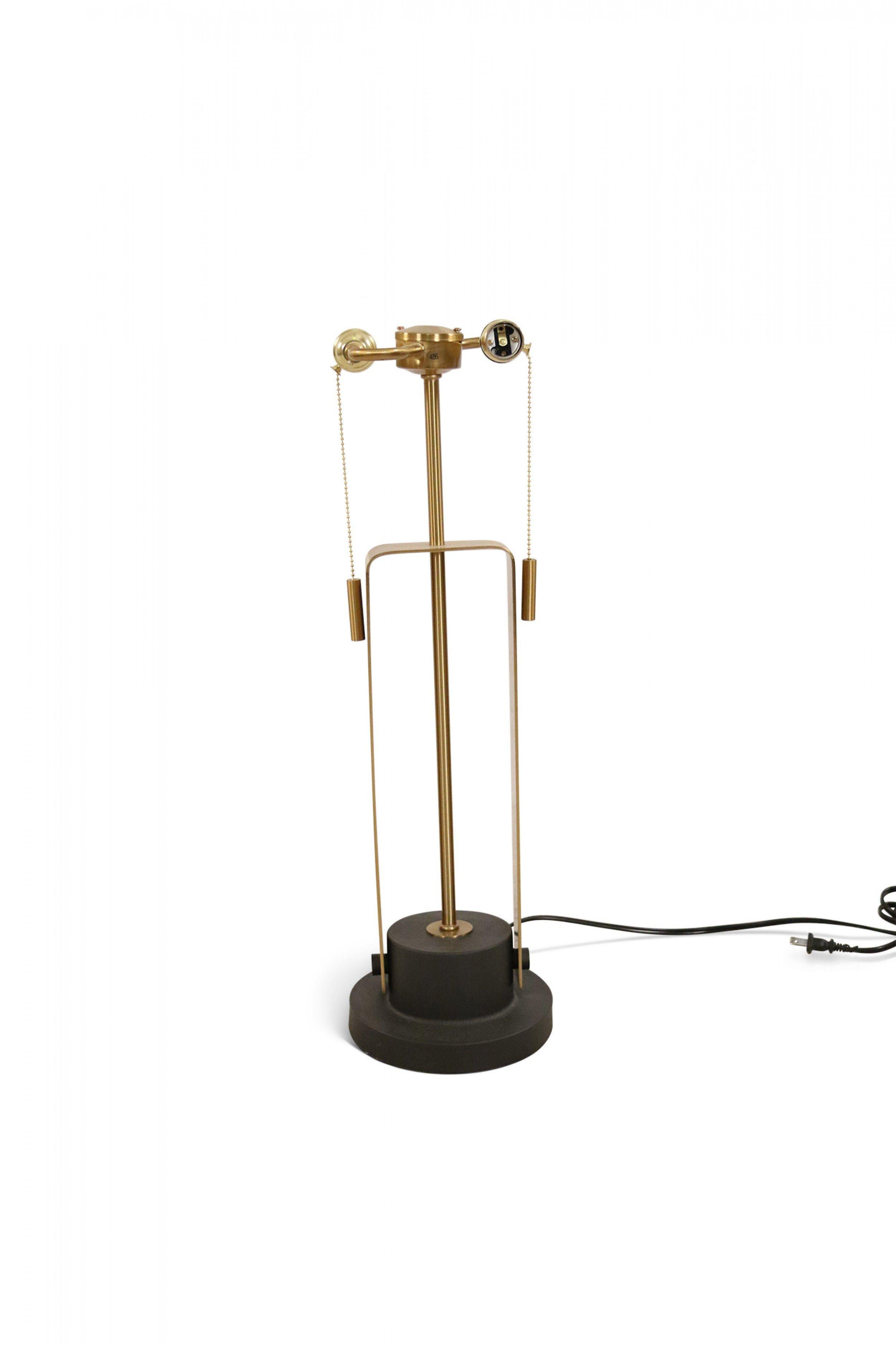 Contemporary polished brass table lamp with a beige fabric drum shade inside a geometric design brass frame on a tiered black metal base with brass supports.