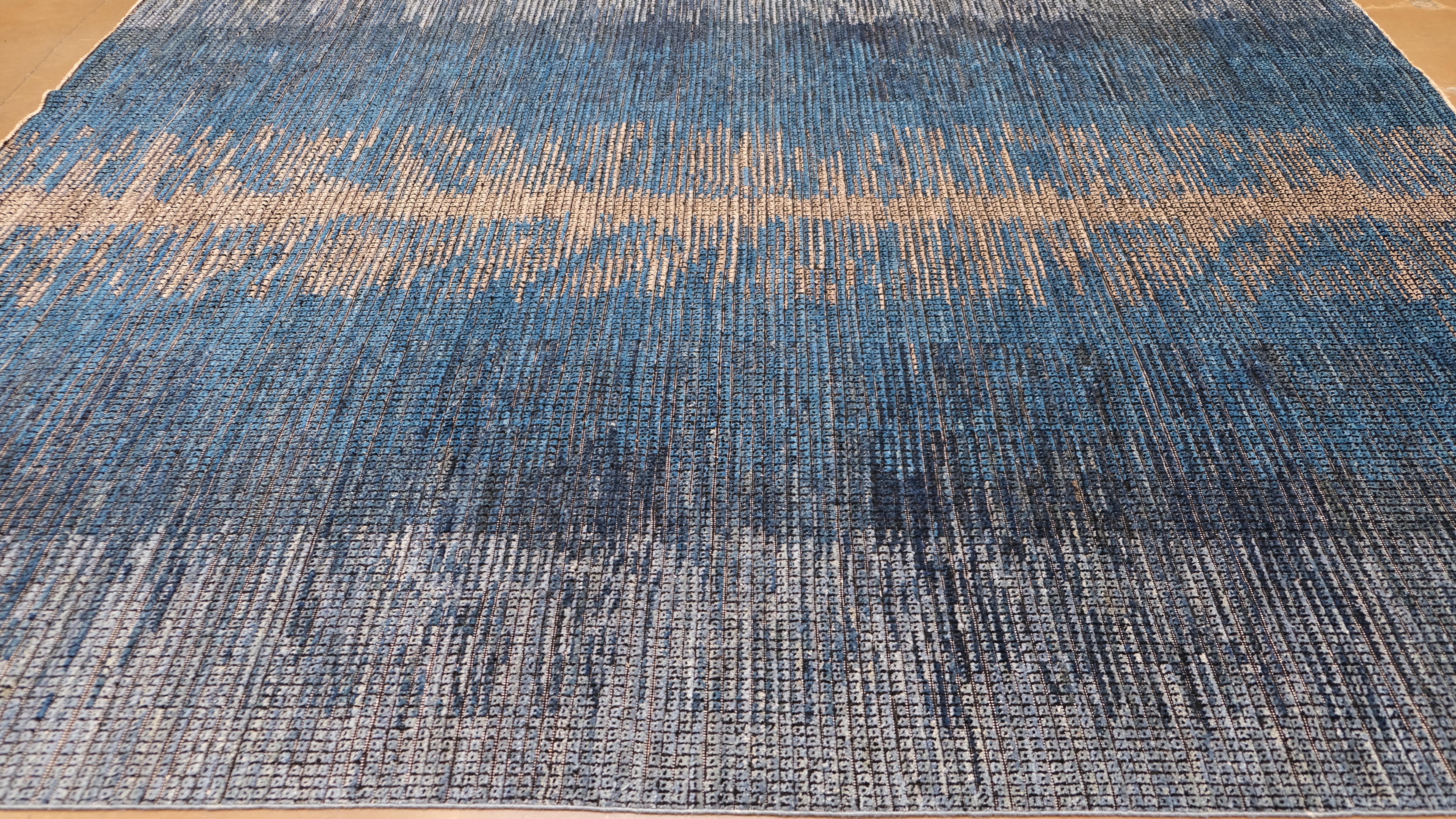 This hand-knotted wool rug is a stunning piece that blends contemporary artistry with traditional craftsmanship. Featuring a bold vertical beige abstraction against a deep blue and gray gradient background, this design evokes the organic beauty of
