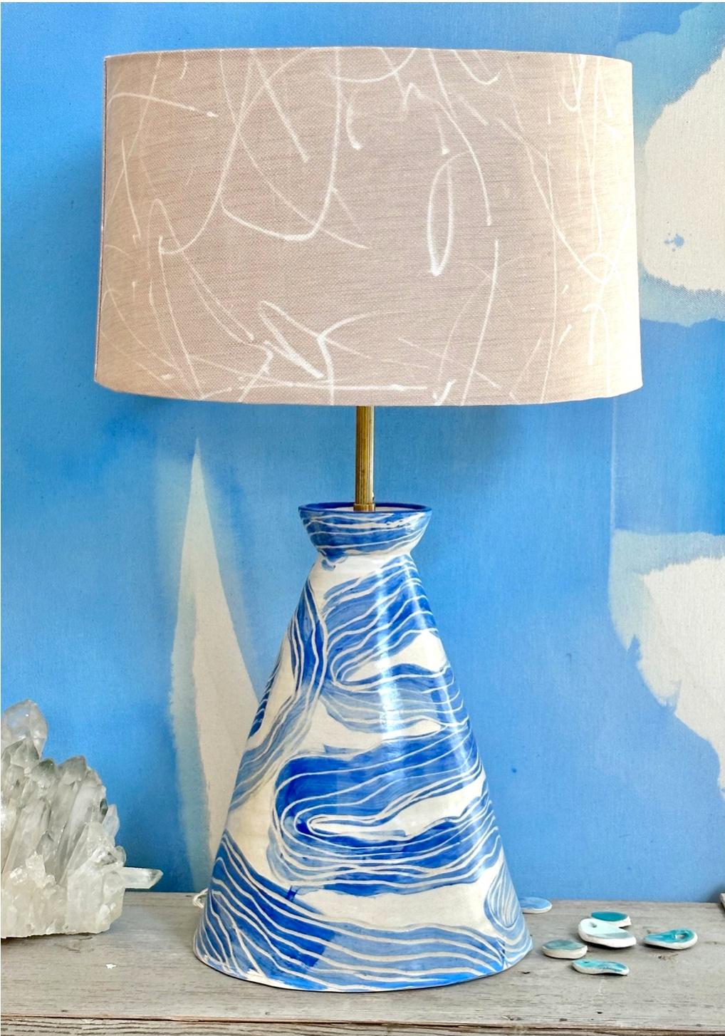 White painted stoneware with a blue painted and incised ribbon

Stoneware ,live brass, off white twisted cord,  shown in custom shade in Scribble by artist Kiki Slaughter

Ceramic height is aprox 13” tall, 9” w, 19.5” to the base of the bulb 
Shade