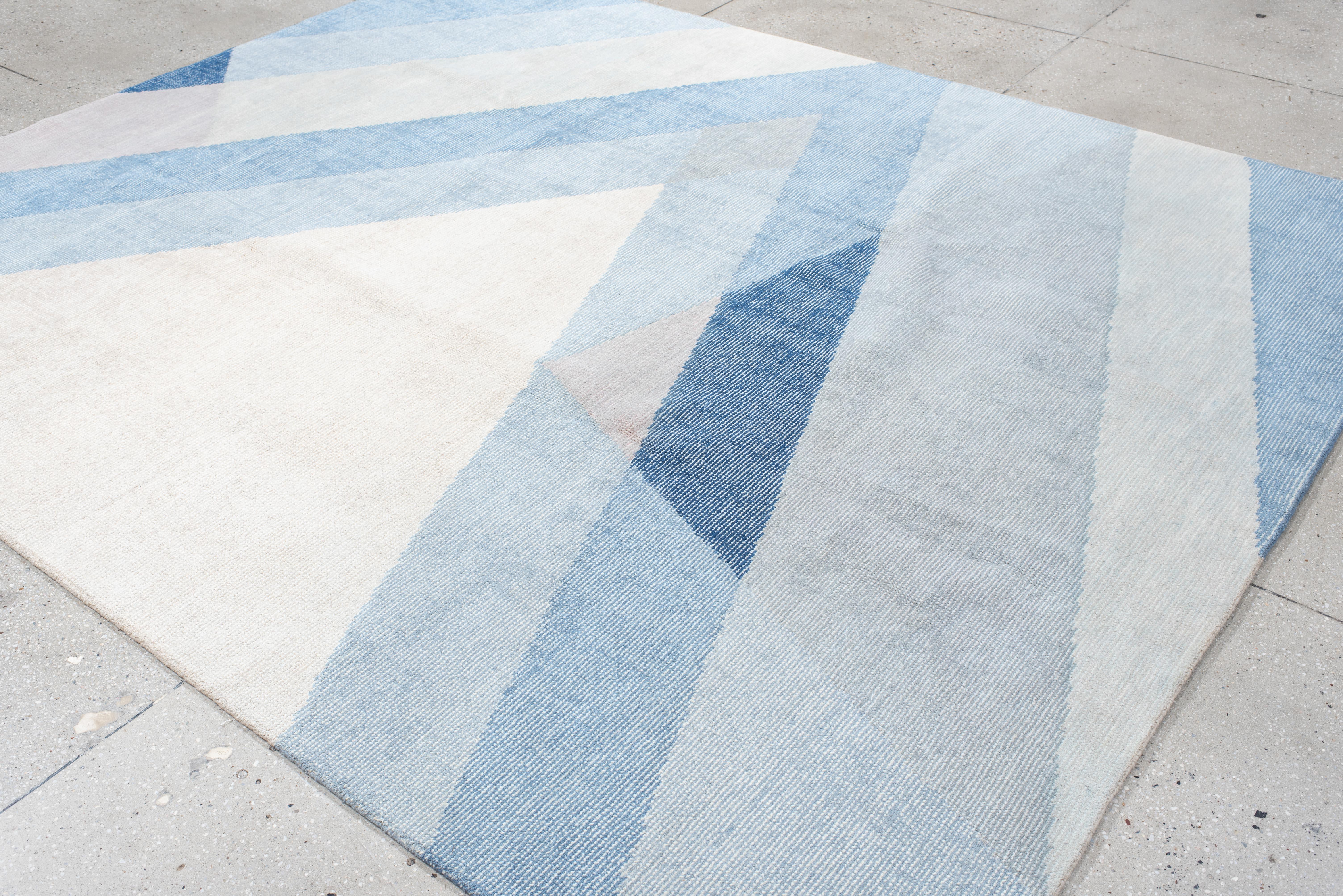 This contemporary hand-loomed rug features a striking arrangement of diagonal bands and angular forms in layered shades of ice blue, pale lavender, soft ivory, and deep navy. The design creates a visual sense of forward movement and crystalline