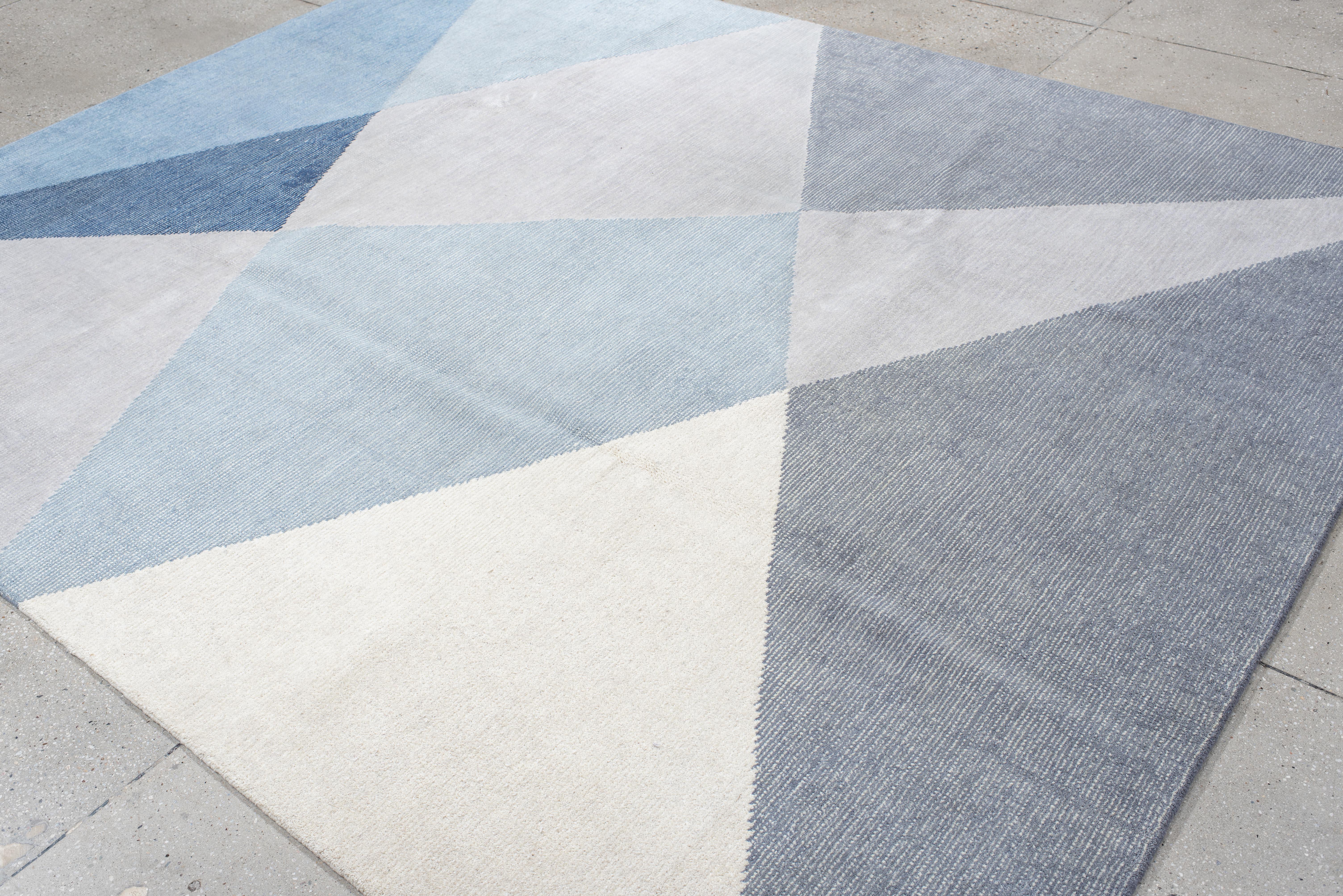 Ce tapis contemporain tissé à la main présente un design géométrique net composé de grandes facettes triangulaires qui s'entrecroisent, dans des tons doux de bleu poudre, d'ardoise, d'ivoire et d'acier. La composition angulaire ajoute une qualité
