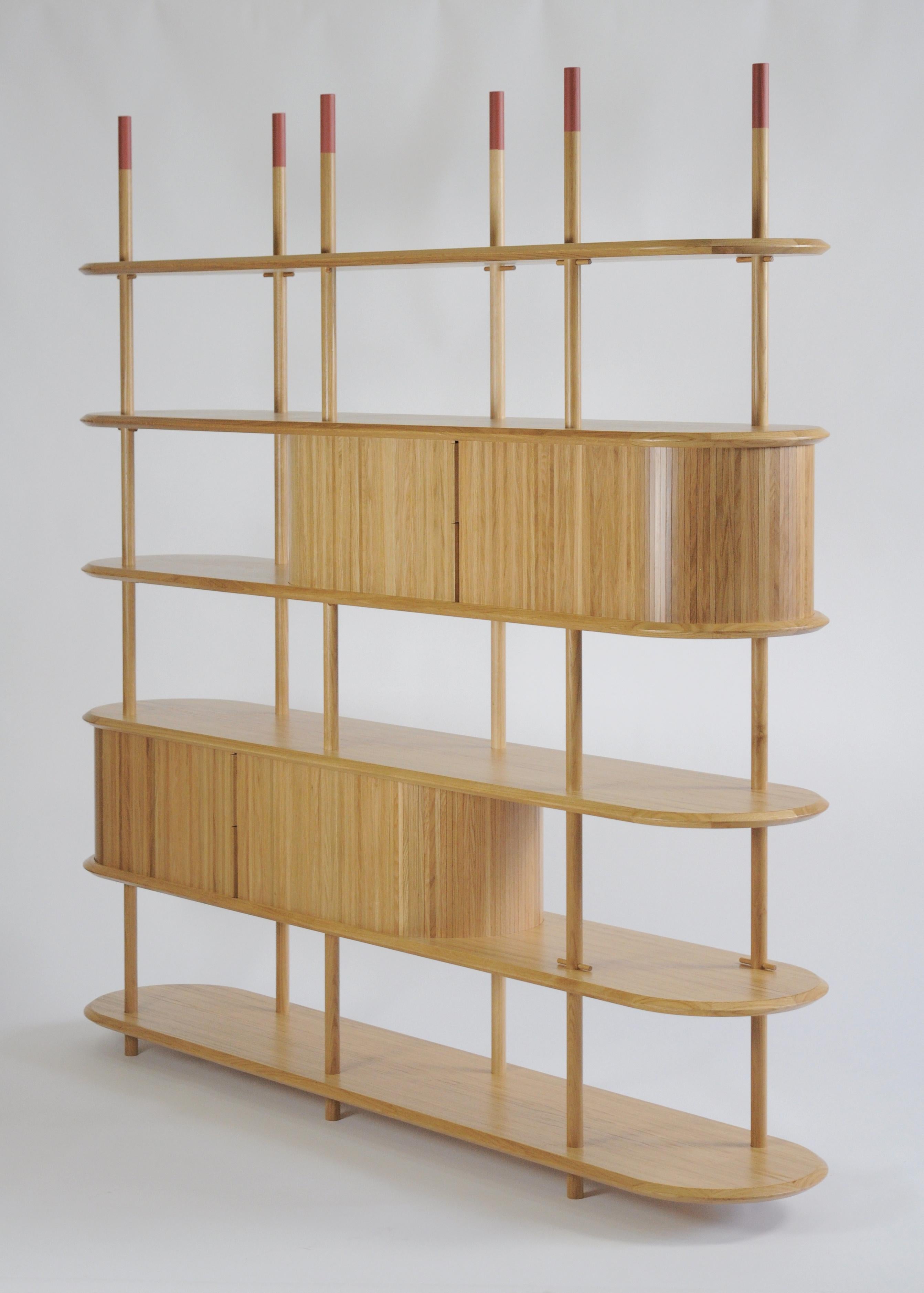Bookcase made of a series of solid wood poles with circular section, veneered shelves with solid wood edging, all made of Oak wood finished with transparent paint. The head of the poles is coated with a brick red protective paint.
The system is