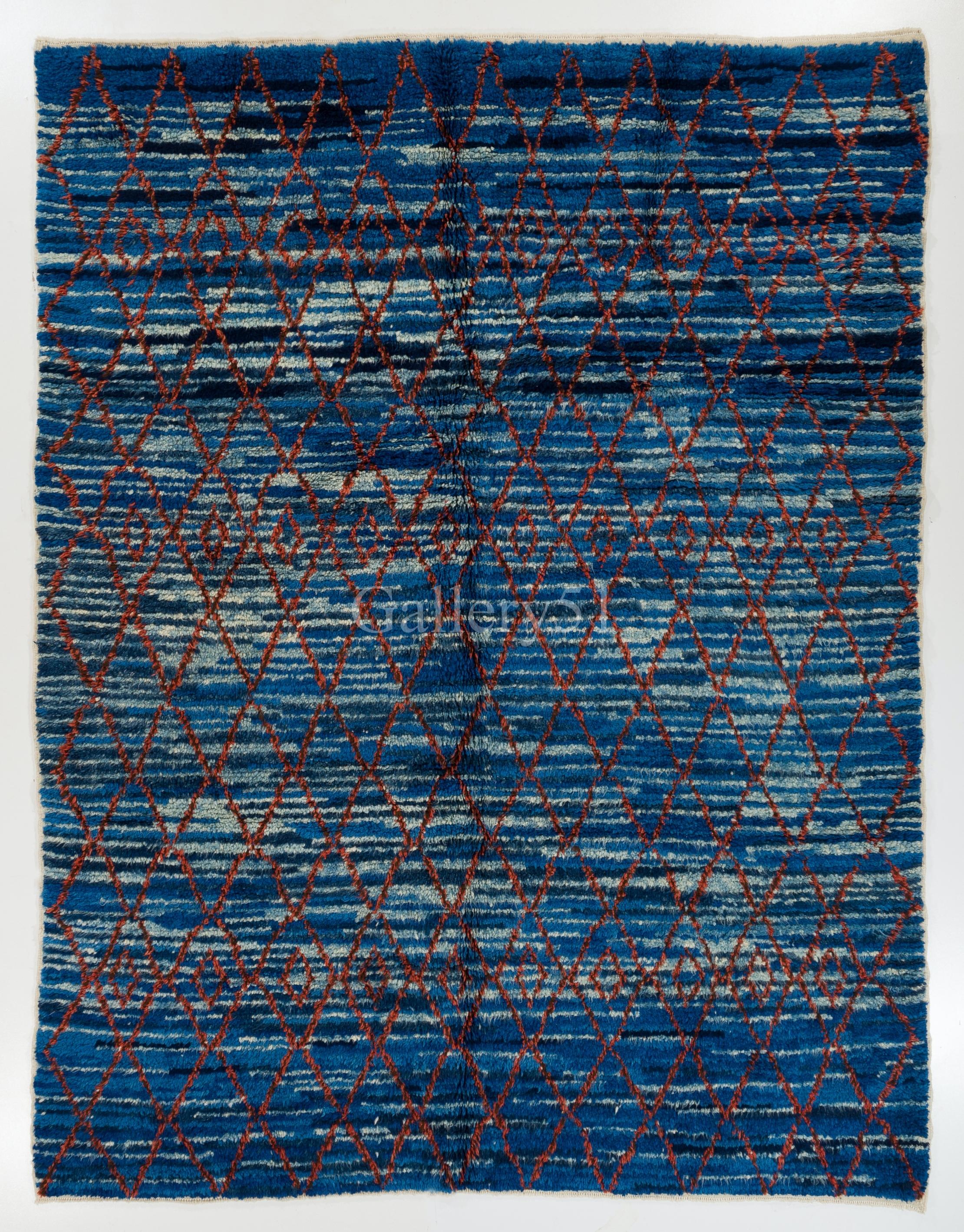 Ein moderner handgeknüpfter Teppich aus organischer Wolle mit marokkanischem Design, der ins Auge fällt. Es schimmert in dunkleren und helleren Blautönen mit einem roten Netz darüber.

Dieser wunderschön gestaltete Plüschteppich besteht vollständig
