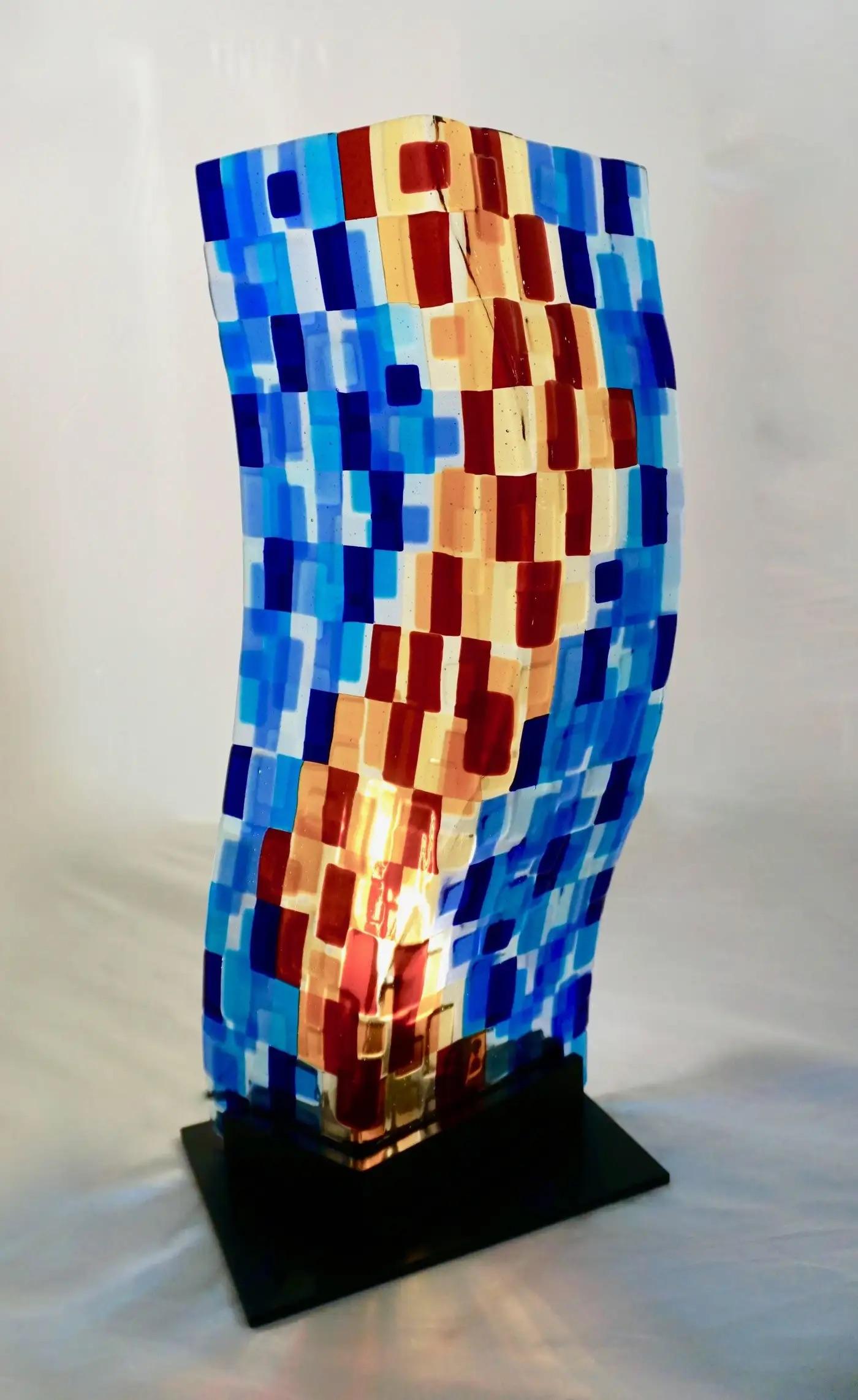 Zeitgenössische, moderne, abstrakte Skulptur aus Murano-Glas, eine dekorative, beleuchtete, gebogene Glasplatte auf einem schwarz lackierten Metallsockel, realisiert als farbenfrohes Mosaik in Azur- und Blautönen, Gelb, Rot und Burgunderpflaume: