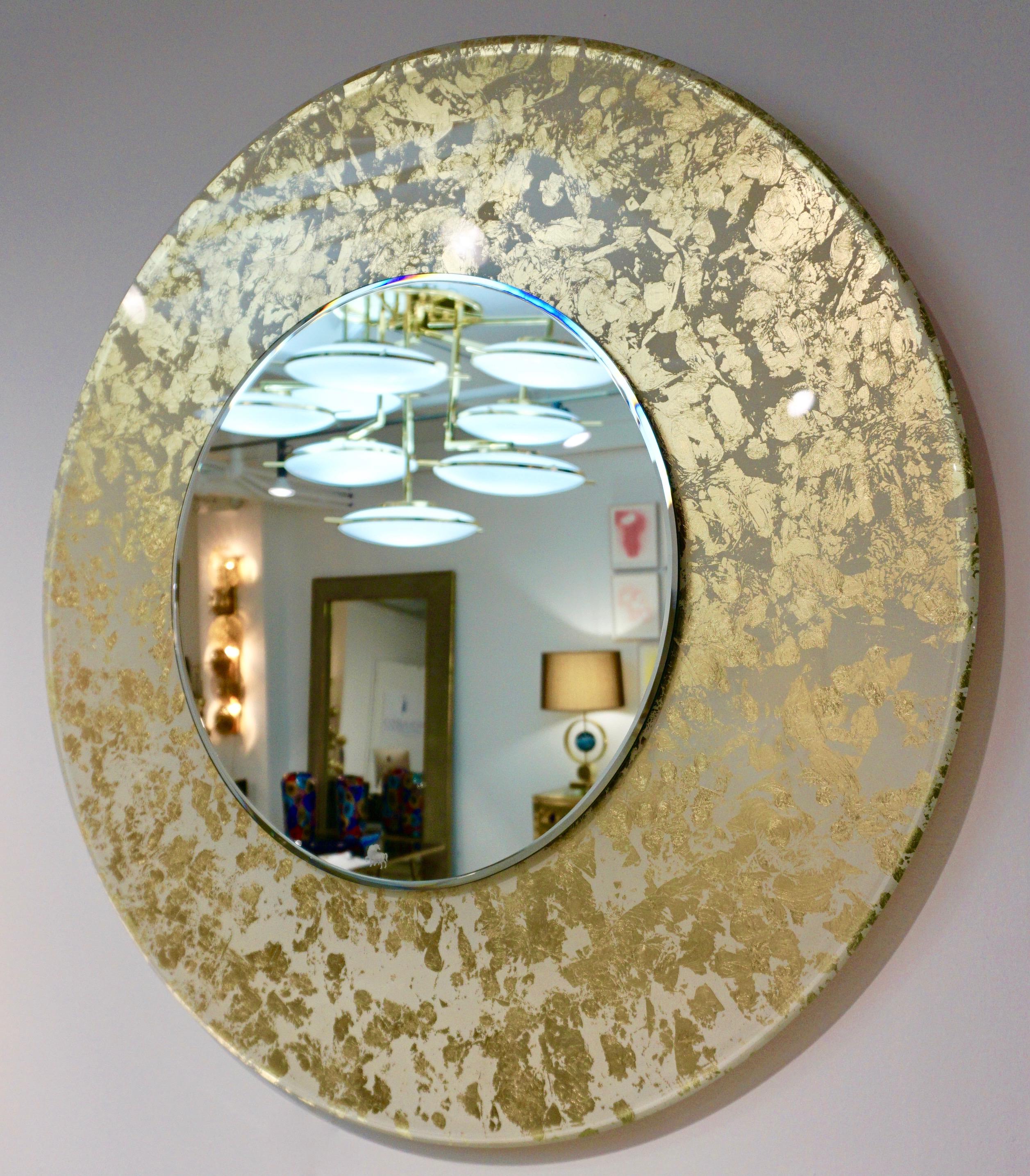 A modern Made in Italy round mirror, entirely handcrafted in glass, of high quality execution: the thick glass frame with beveled edge is decorated on reverse in cream white and a pure gold leaf blotch design that glows when illuminated. Fitted with