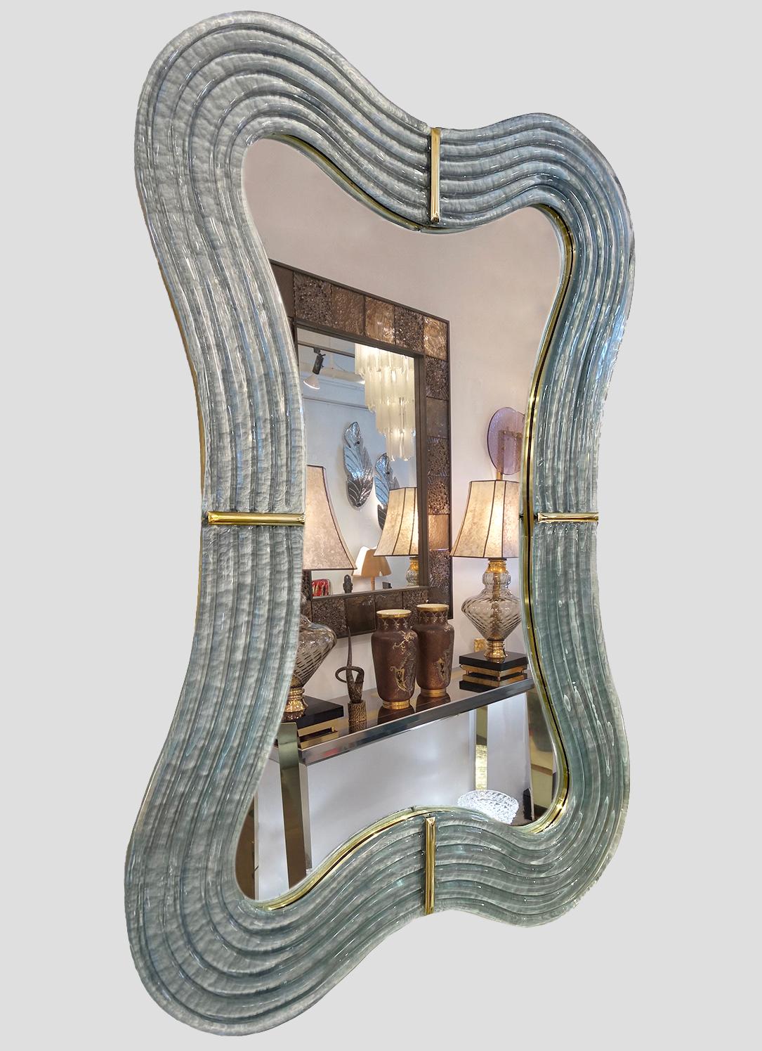 This Venetian mirror brings a sensuous soothing feeling with its curved silhouette, entirely handcrafted in Italy. The sexy waved shape presents exquisite craftsmanship: the hand-carved wooden back, entirely edged in brass, is decorated with a