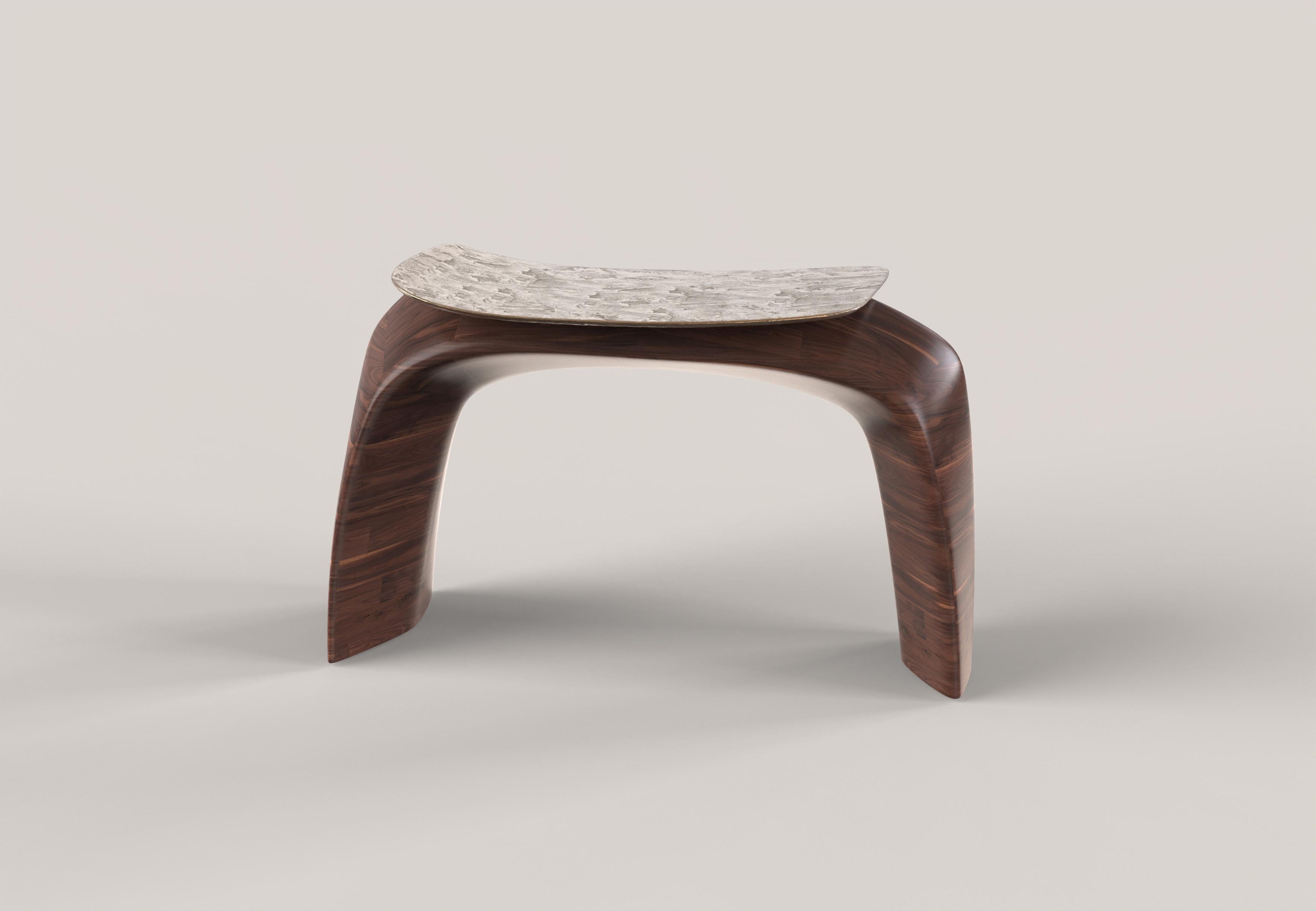Ce tabouret du XXIe siècle est conçu en travaillant avec une approche sculpturale sur deux matériaux : le bois massif et le bronze coulé. Le bois est sculpté, créant un arc de lignes à la fois douces et fermes qui constituent la structure de