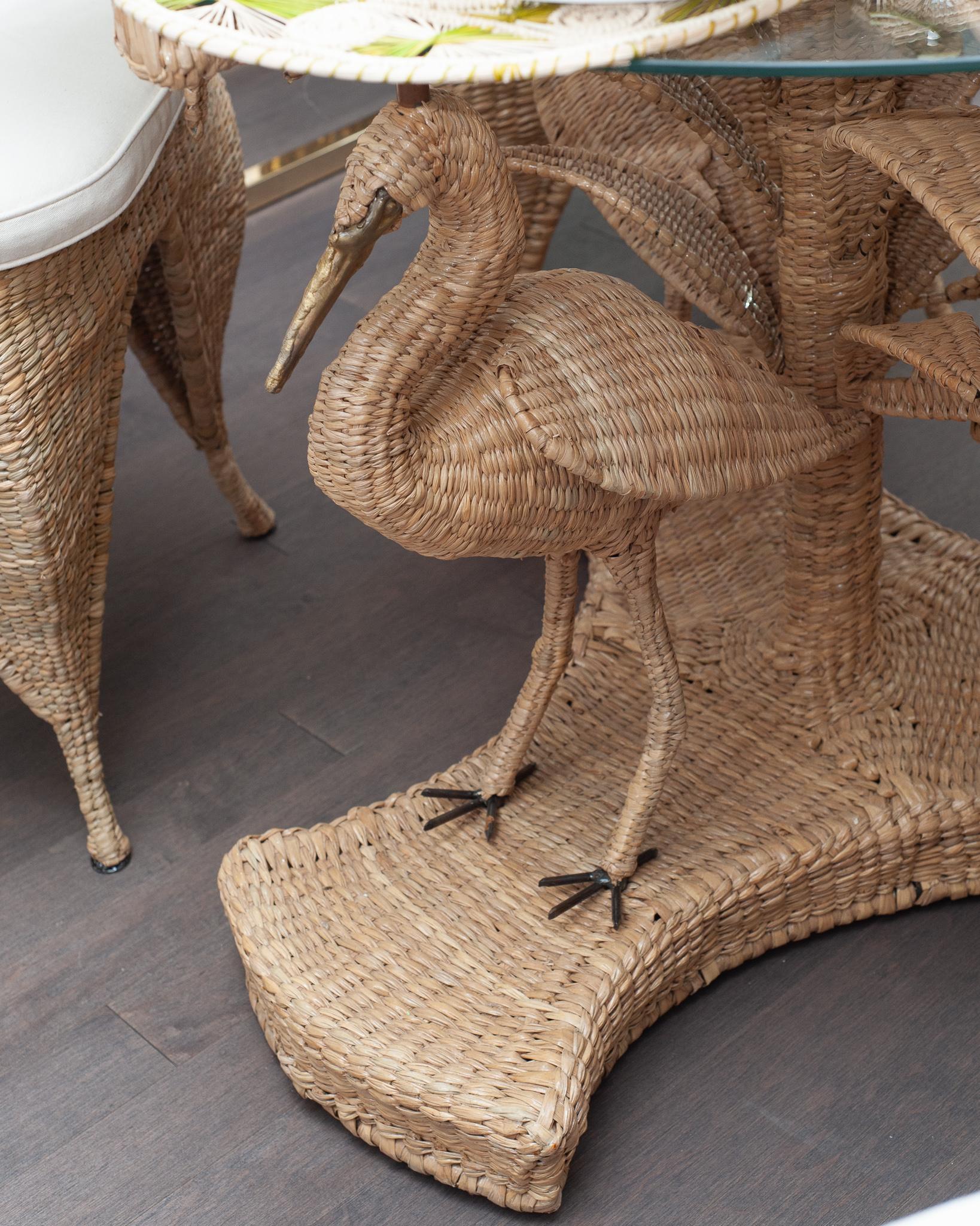 A contemporary Mario Torres large crane table, constructed with a metal frame supporting a glass top and expertly hand woven natural rattan. This table features palm leaves and cranes with pressed brass faces and beaks. Labeled with a brass