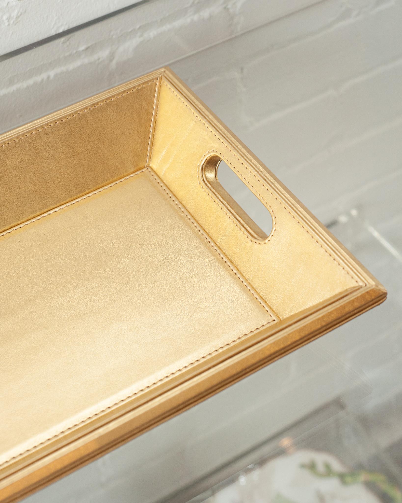 A beautiful metallic gold leather tray, perfect for the desk, bedside table, or use as a serving piece. Fully wrapped in embossed leather with stitched panels, each tray has two openings for carrying.