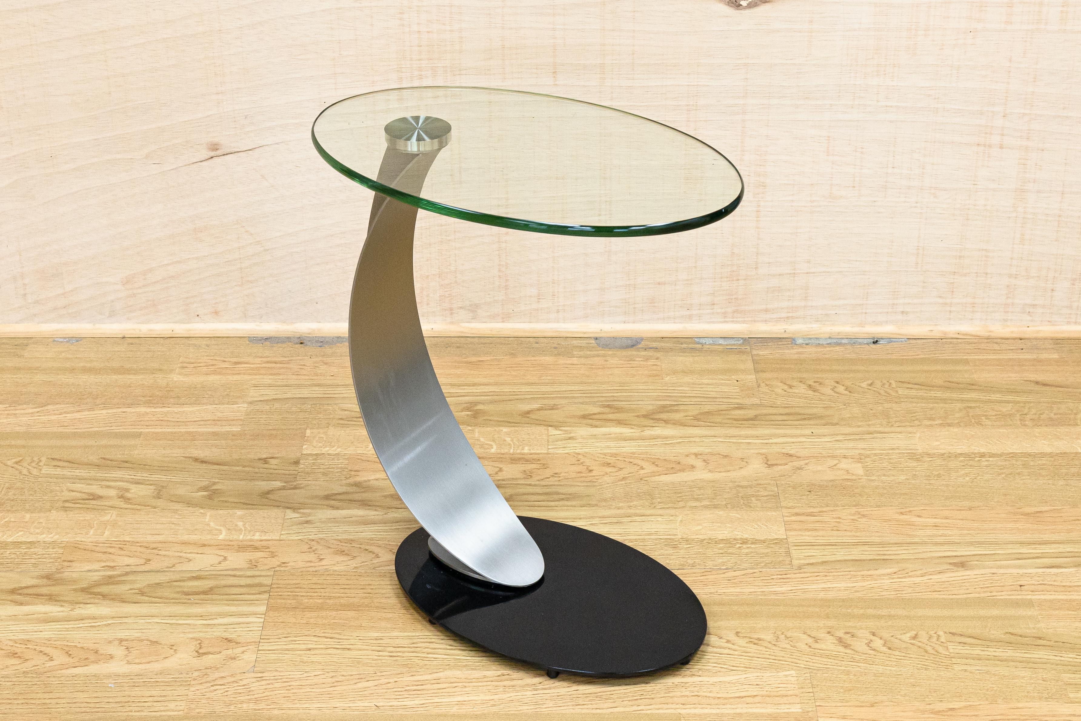 A DIA polished chrome and glass curved side end table. This elegant side/end table features a clean oval glass top with a dynamic curved metal frame leading down to a heavy black base. This piece is in vey good vintage condition. It measures 21.25