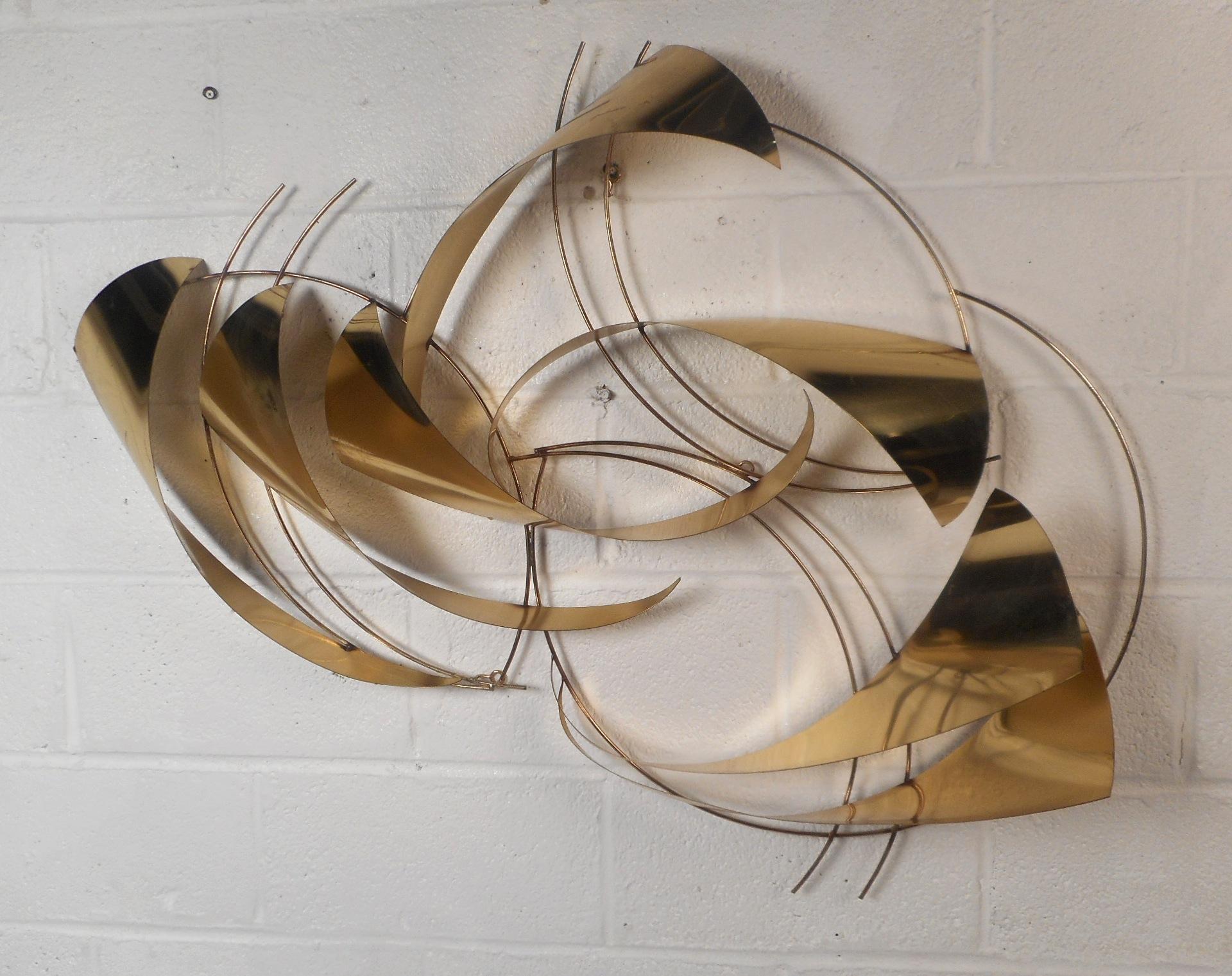 This unique midcentury style metal wall art features a stunning three dimensional design. The item is comprised of multiple sculpted shapes and an elegant golden color. This fabulous wall art can be hung up both vertically and horizontally, giving