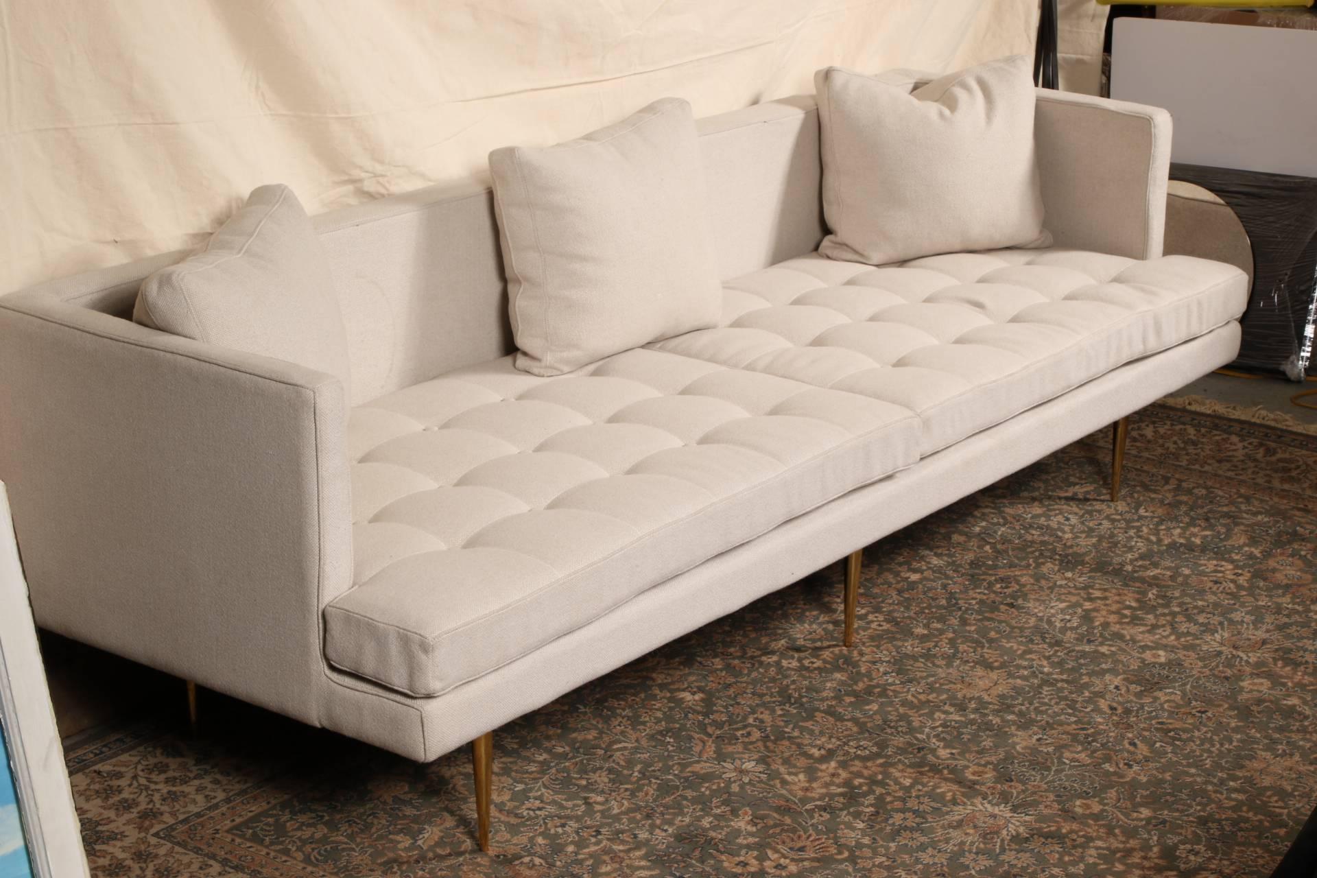 Contemporary modern sofa, white upholstery with a two part button- tufted seat and vertical arms, raised on tall cylindrical tapering brass legs, accompanied with three toss pillows, missing the back cushions.

Condition: Expected wear and signs