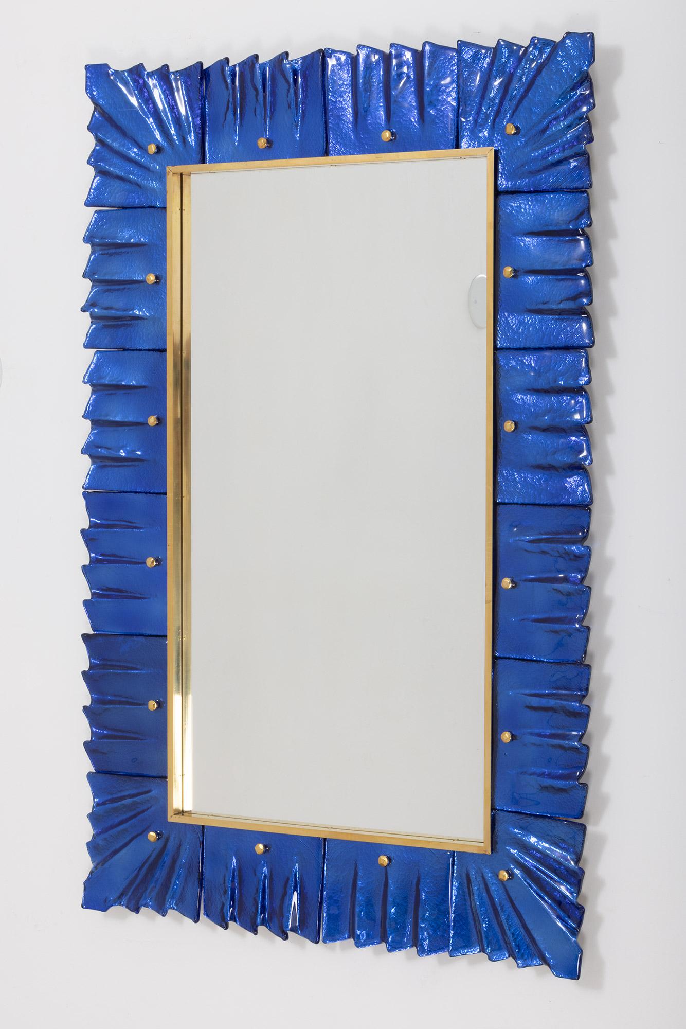 Miroir contemporain encadré en verre bleu cobalt de Murano, en stock
Plaque de miroir entourée de carreaux de verre ondulés de couleur bleu cobalt retenus par des cabochons en laiton. 
Fabriqué à la main par une équipe d'artisans à Venise, en