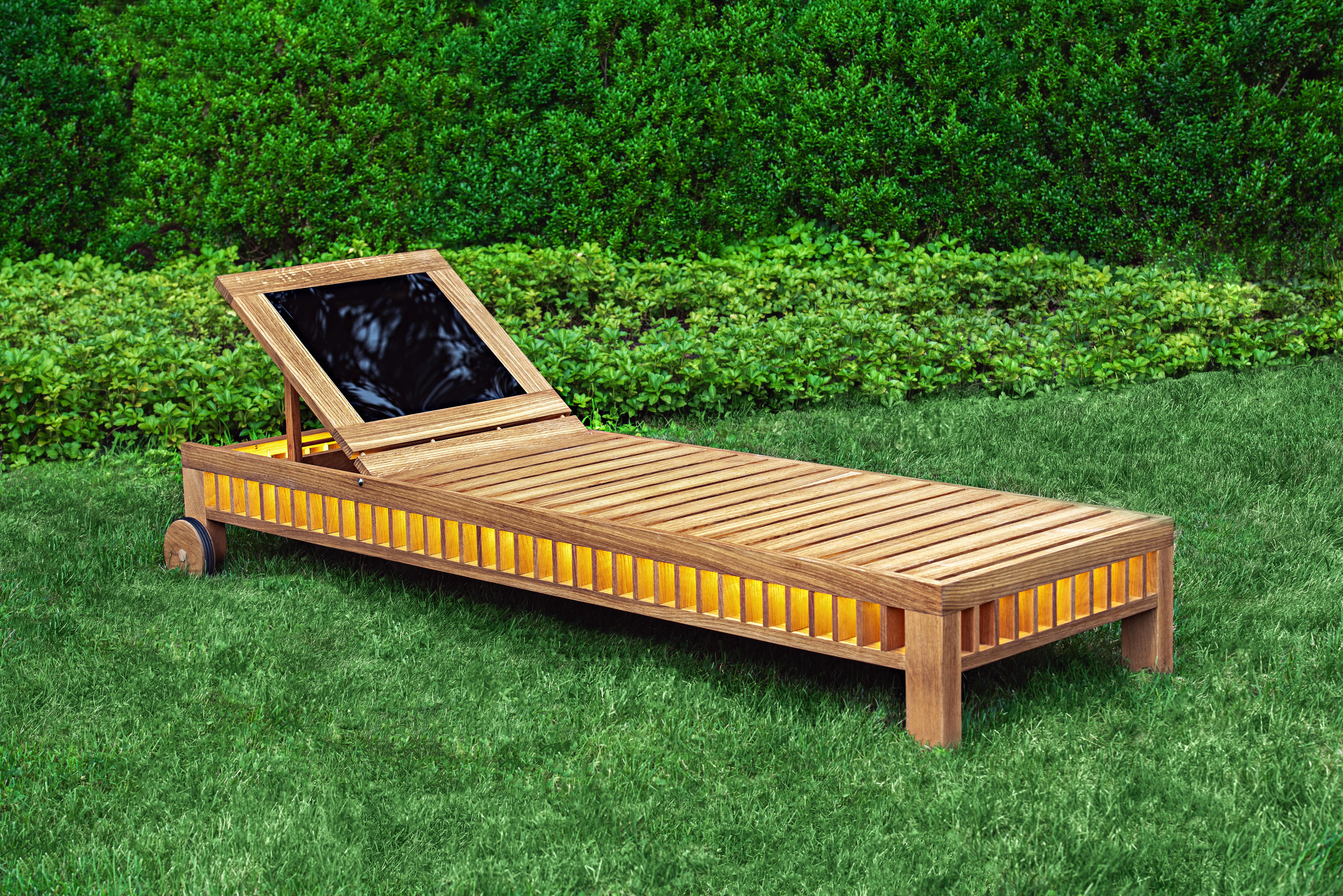 This wooden slatted solar lounger integrates a photovoltaic panel in its adjustable backrest. The panel, which reflects the surroundings in its mirrored surface, collects enough power to illuminate lights inside the body of the lounger at fall of
