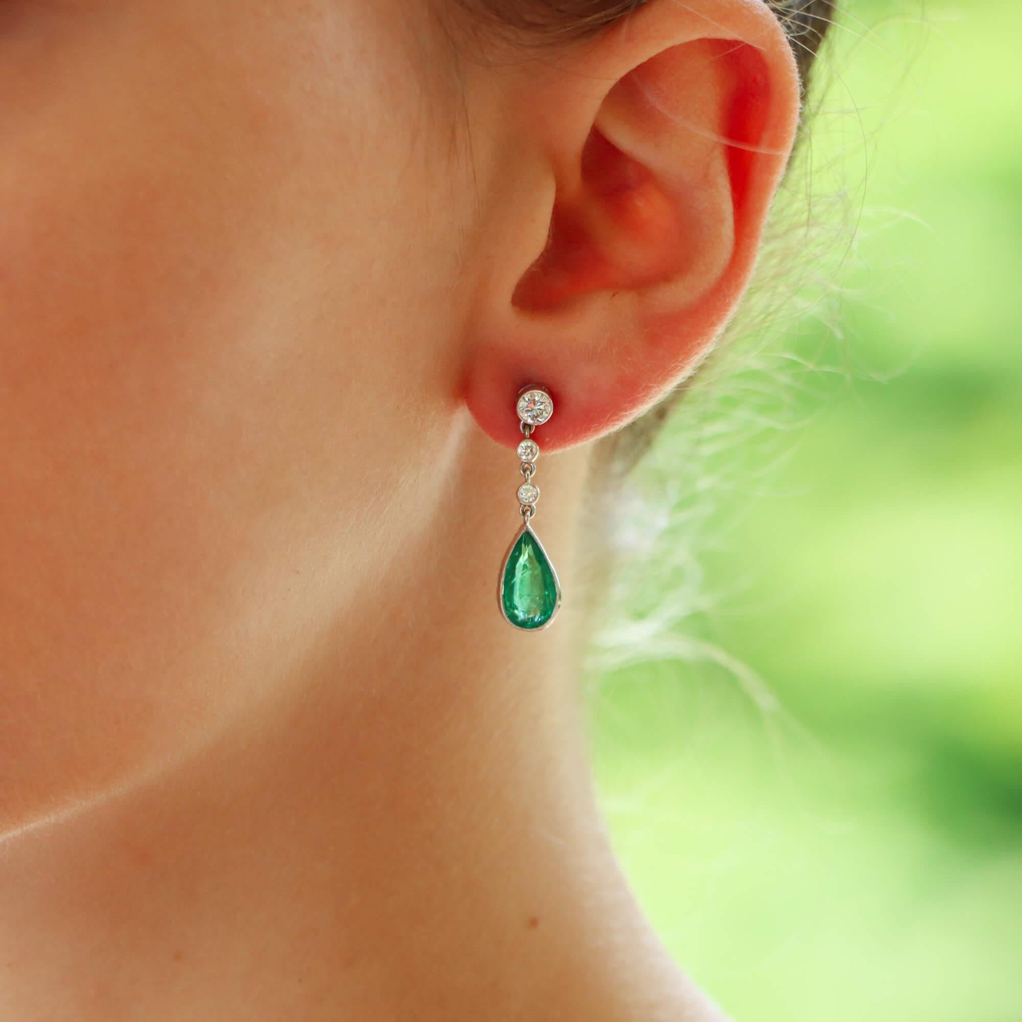 A beautiful contemporary pair of emerald and diamond drop earrings set in 18k white gold. 

Each earring prominently features vibrant pear cut emerald stone that elegantly hangs in a rub over setting. To the top of the pear cut emerald is a