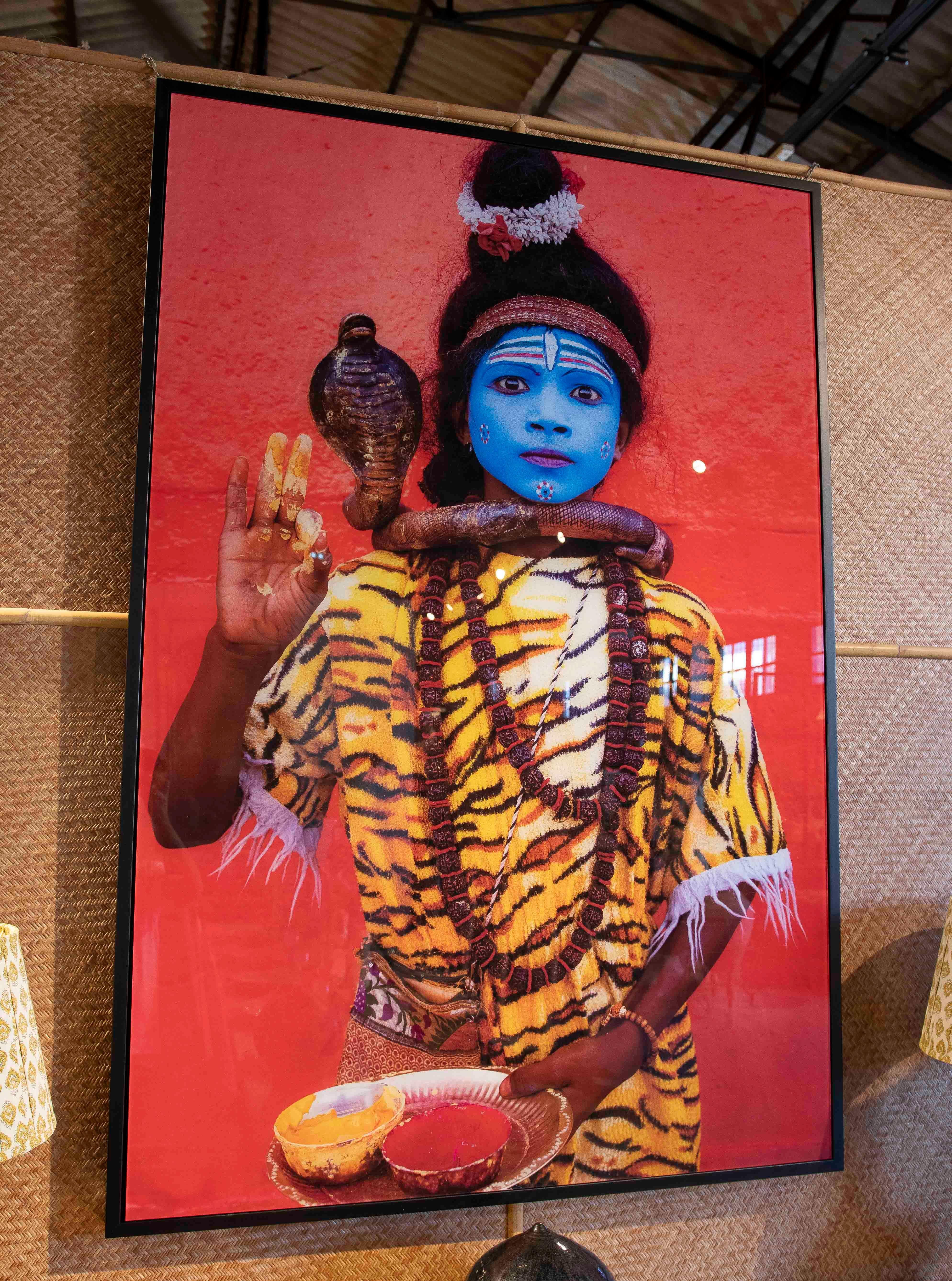 A stunning contemporary photograph depicting a young man dressed as the Hindu god Shiva, with his face painted blue, a snake around his neck, and a tiger-skin garment. The portrait conveys the spiritual and symbolic power of one of the most revered