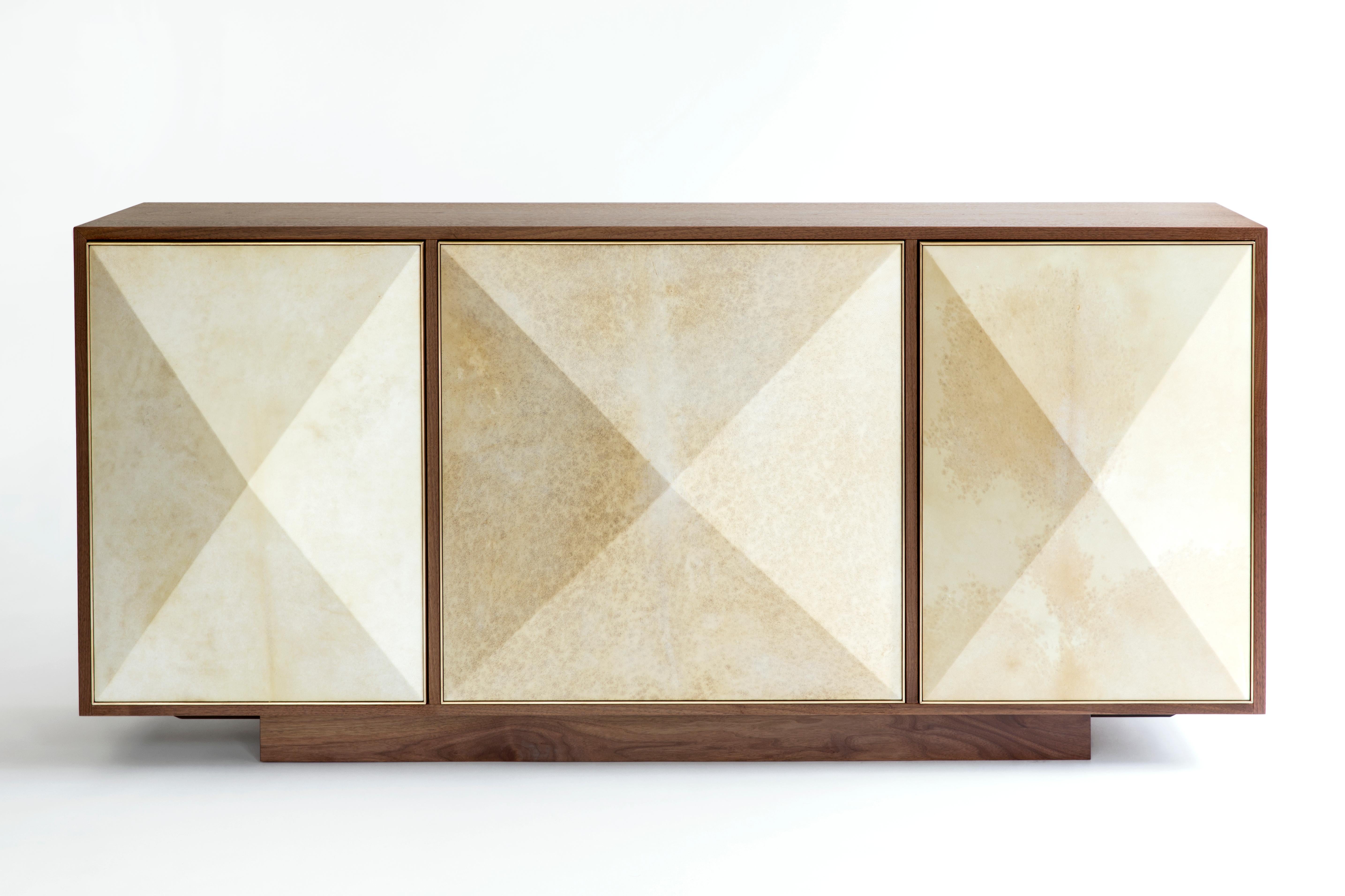 The 3 door Pyramid Sideboard features three three-dimensional doors hand-wrapped in natural goatskin parchment and framed subtly in satin brass. The recessed base gives the piece a floating appearance. The outer doors of the sideboard are slightly