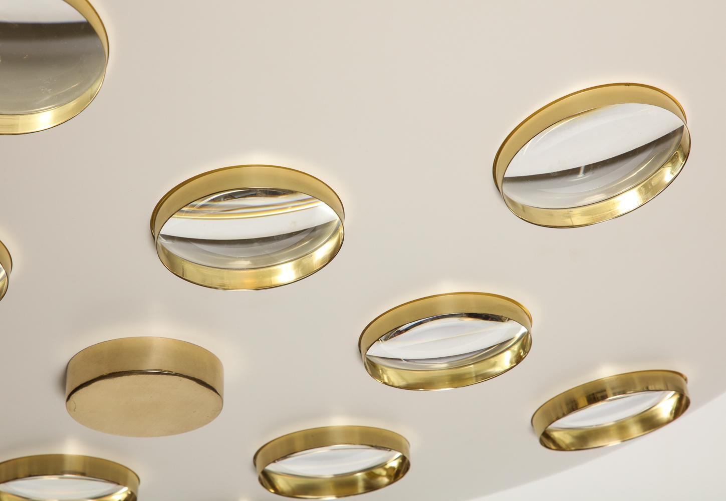 Beautiful cream-painted aluminum saucer form, with circular cut-outs. Each cut-out is fitted with a brass framed glass lens, creating a subtle abstract effect. 6 standard E26 Edison sockets and polished brass mounts. The rod can be cut down and this