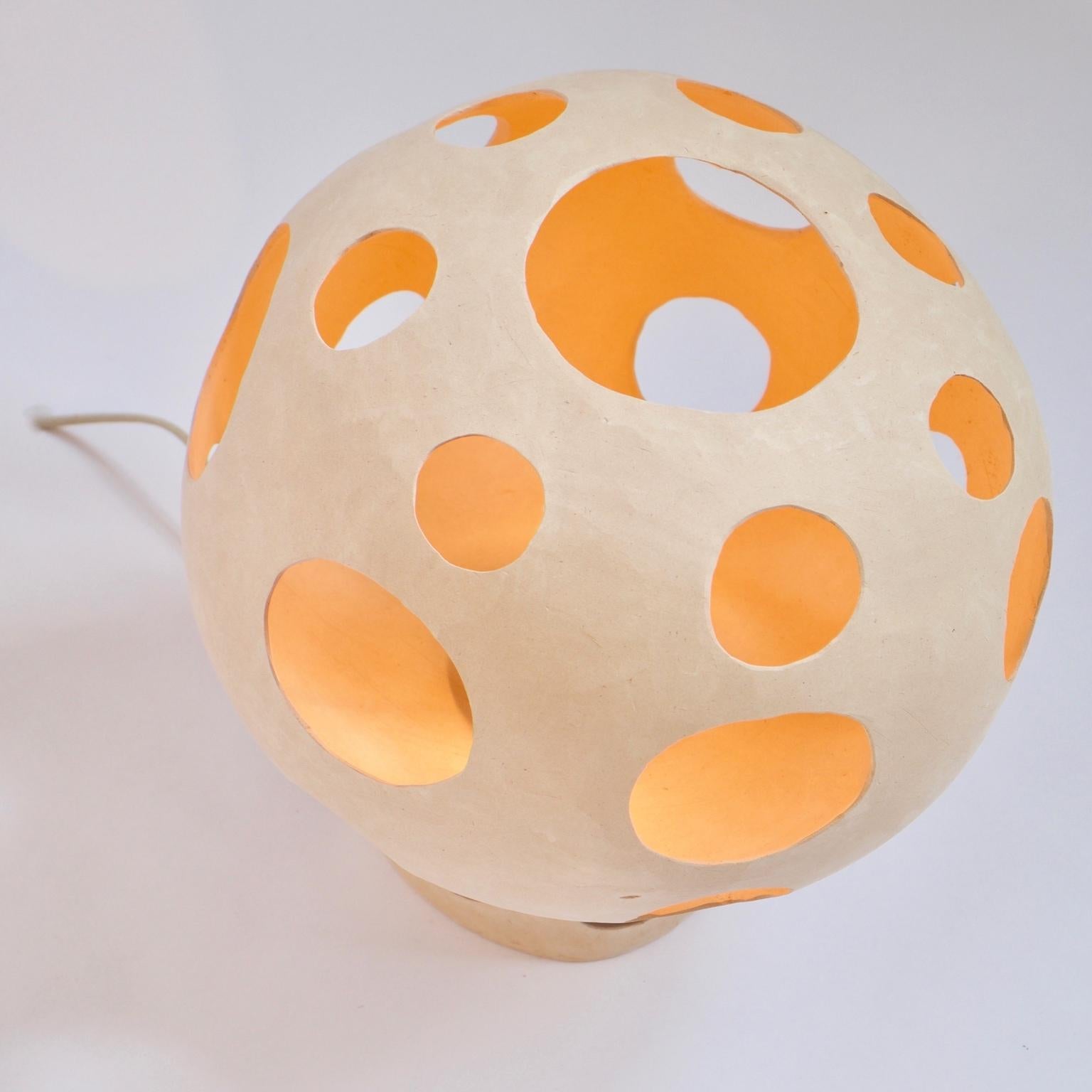 Persimmon is a mushroom-shaped hand-built ceramic table lamp whose plinth base and socket cover are integrated into the main body making one seamless sculptural ceramic shell. The cutouts on the body add character and project multiple round spots of