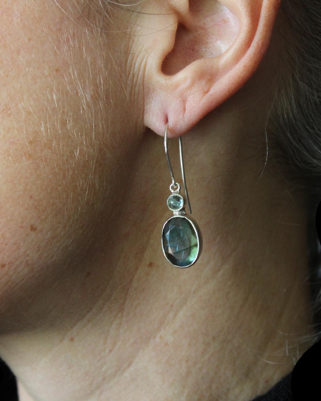 These pretty earrings feature faceted labradorite cabochons complimented by light blue, round aquamarine accent stones. Labradorite is displays an iridescent effect, which is even more pronounced when the stone is faceted. They hang gracefully with