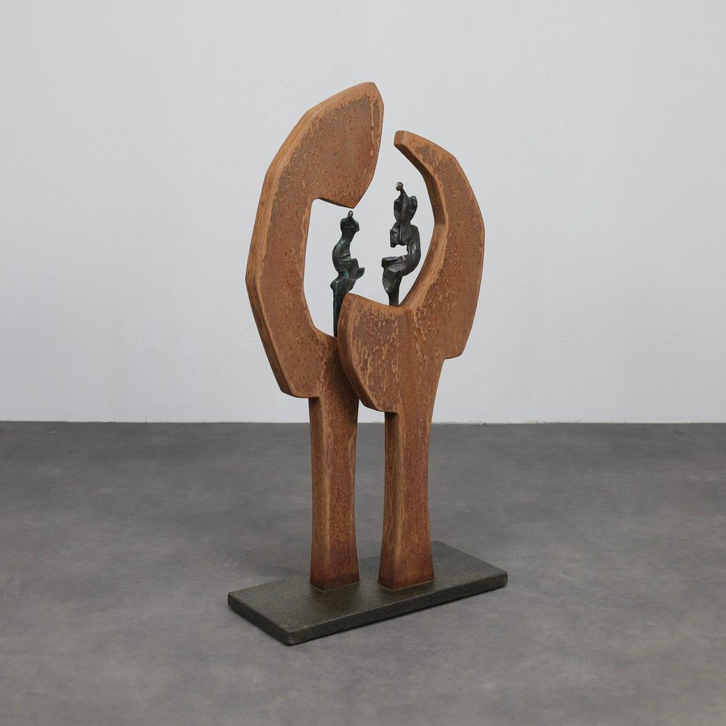 Eine markante und einzigartige Komposition, die rohen Cortenstahl mit zarten Bronzefiguren und einem massiven Steinsockel verbindet. Die Skulptur evoziert sowohl Stärke als auch Intimität: Der verwitterte Stahl bildet eine umarmende Struktur,
