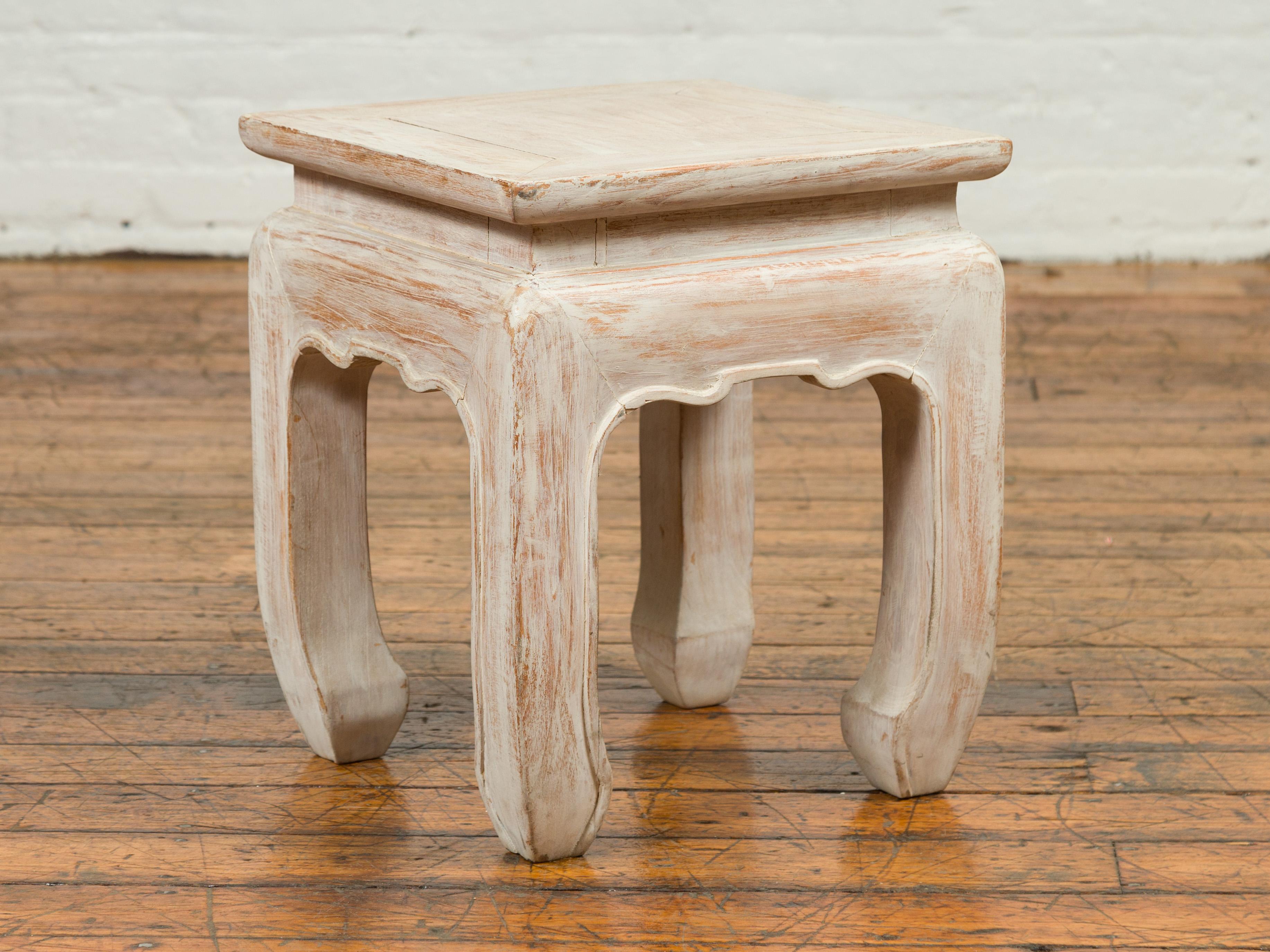 A contemporary Ming style Thai painted teak waisted stool with chow legs and carved apron. Charming us with its elegant lines and distressed finish, this Ming style stool features a square waisted top sitting above four chow legs, connected to one