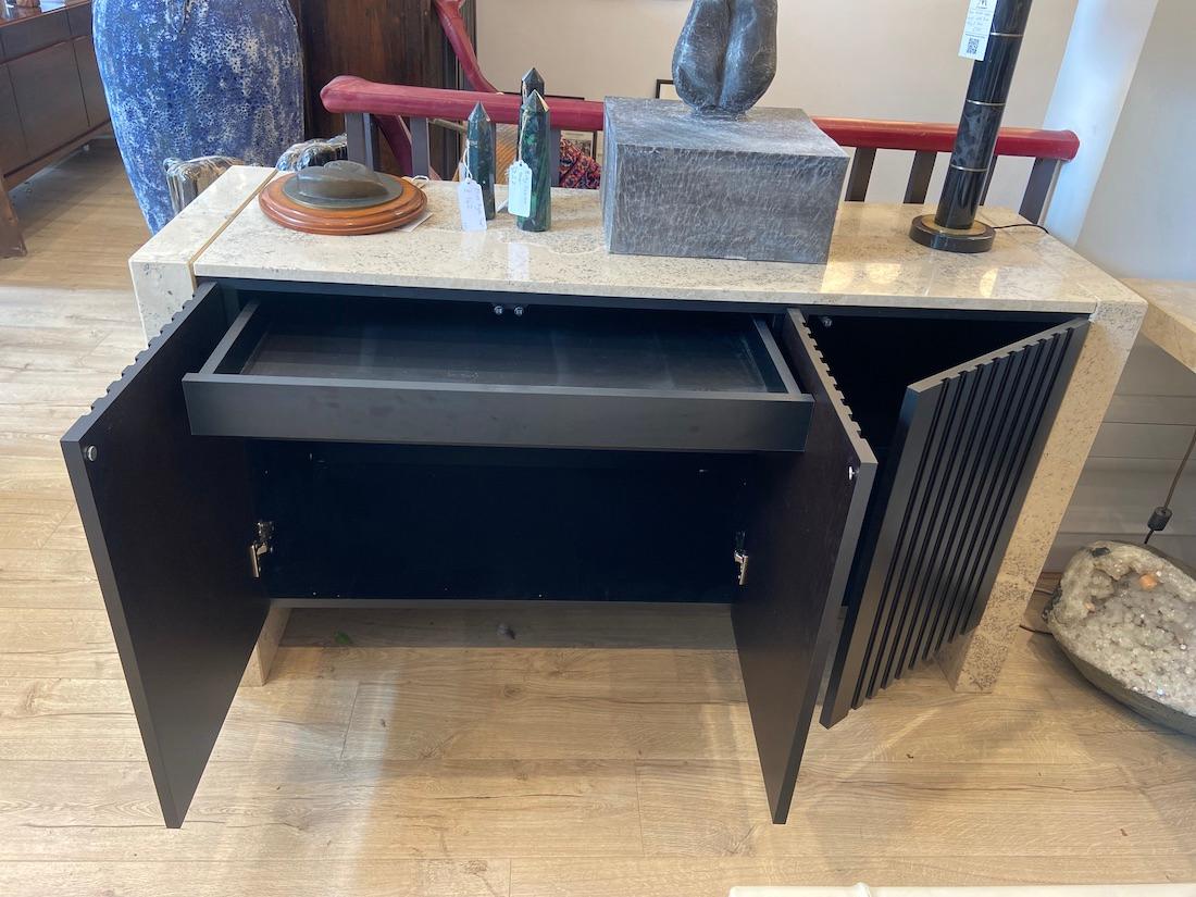Un aparador contemporáneo de travertino con piezas plateadas brillantes. 

Este aparador ofrece un amplio espacio de almacenamiento, con puertas de lamas con frentes negros que se abren a un total de cuatro grandes estantes. 

También hay un cajón
