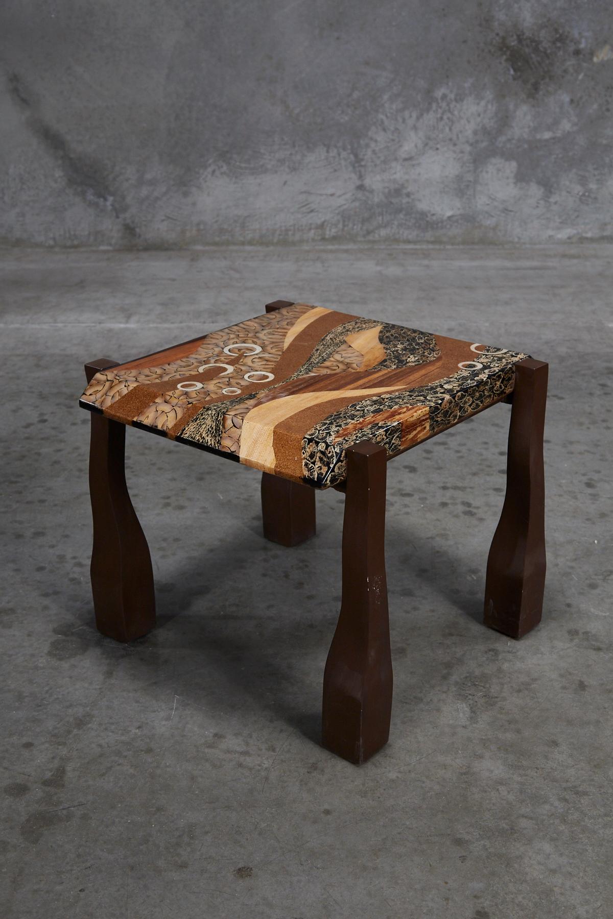 Square side table with exotic inlay over a fiberglass body. Multiple types of materials including wild pearl vine, cotton husk, light and dark banana bark, bamboo circles, and honeycomb cane leaf into a freeform abstract pattern across the surface