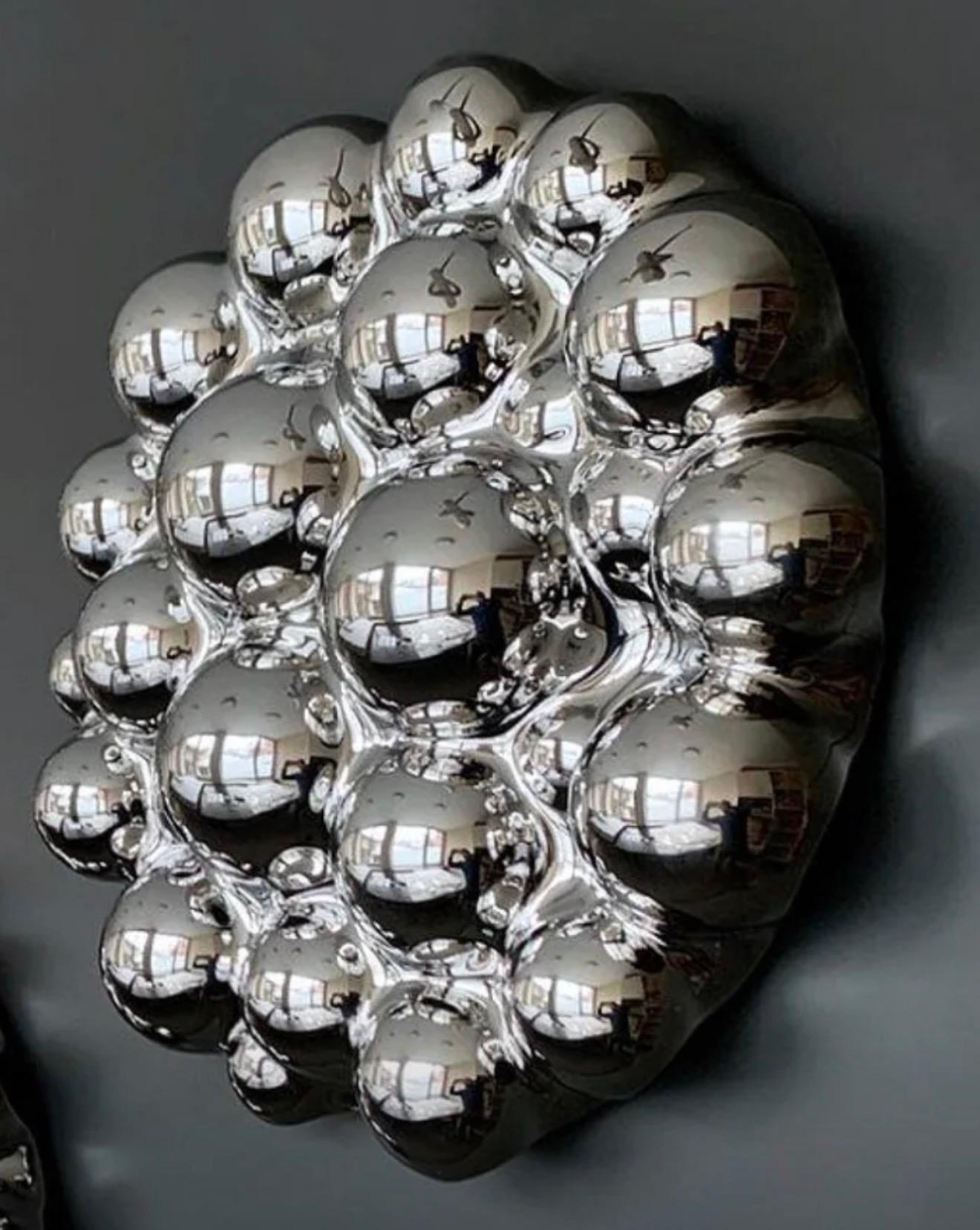 Die silberne Wandskulptur Bubbles besticht durch eine Komposition aus konvexen Spiegelkuppeln, die scheinbar schwerelos an der Wand schweben. Jedes Glaselement hebt sich subtil von der Oberfläche ab und wirft wechselnde Schatten, die sich im Laufe