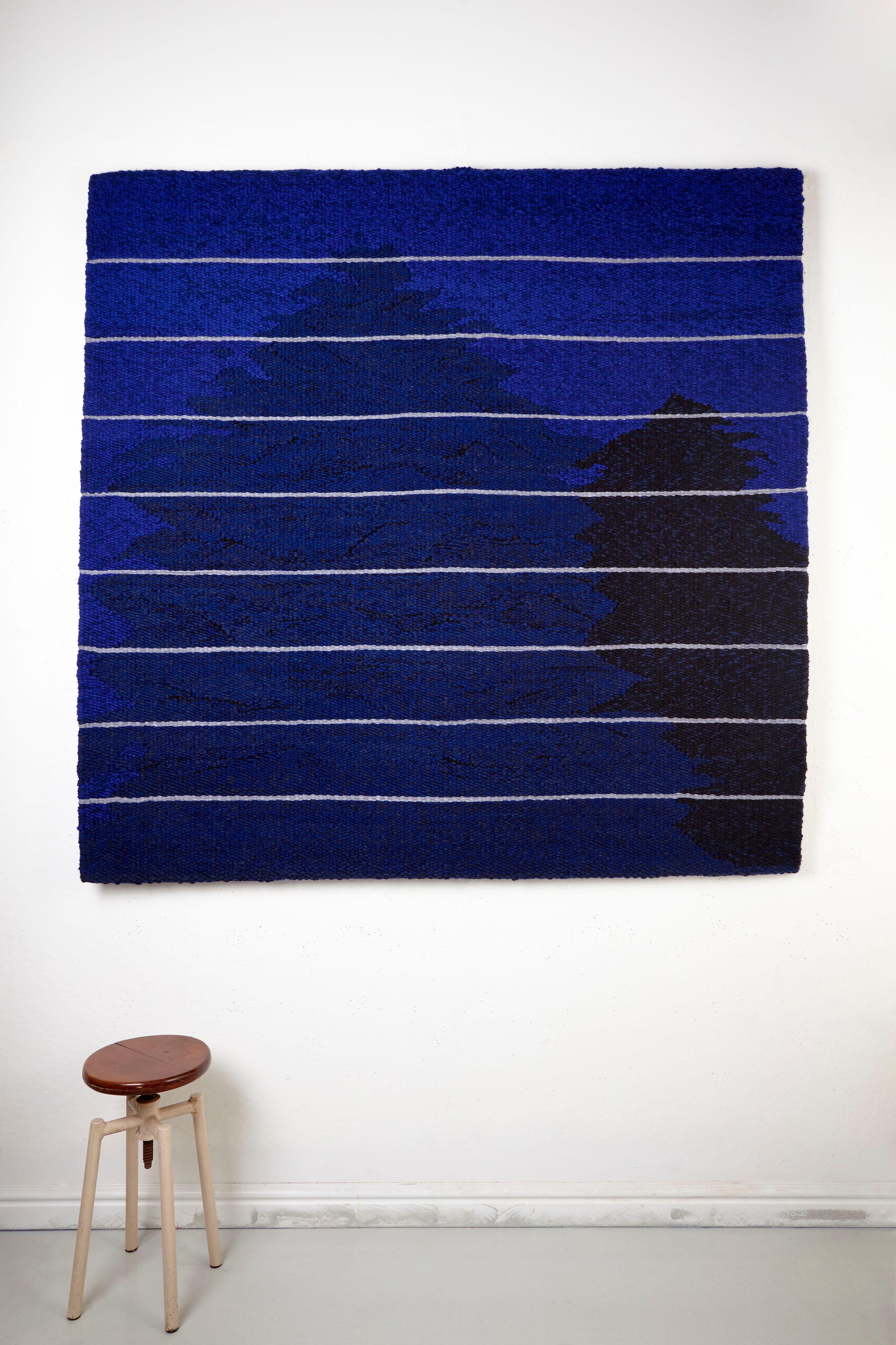 This tapestry titled “Moonlight, midnight and shadows” depicts a moonlit scene with 2 trees silhouetted against a rich blue sky, it is made up of various shades of blue, blue/purple and black, the moonlight is depicted by bands of silver grey across