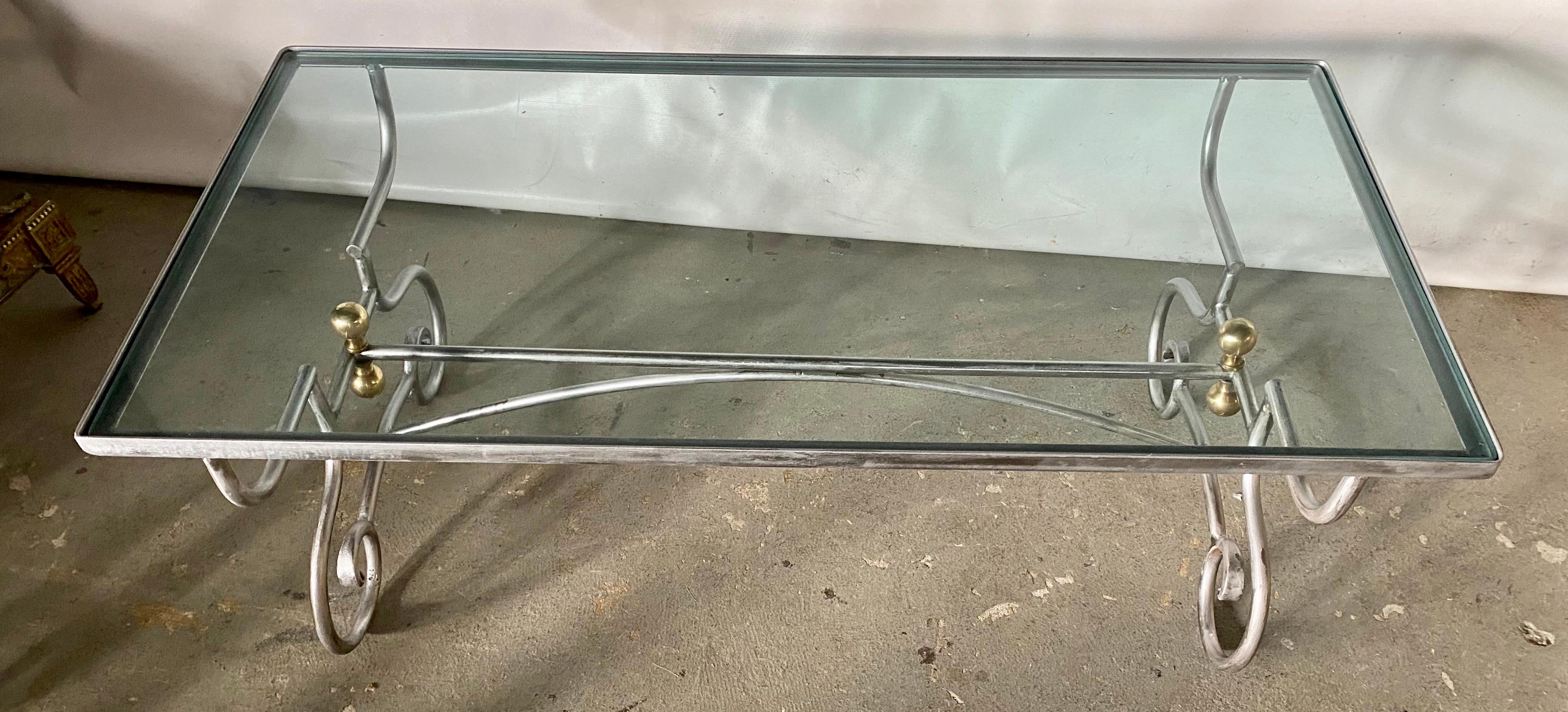 The contemporary Maison Jansen inspired wrought iron steel coffee table has a glass top inset in a frame. The two gracefully curved legs, embellished by pairs of brass knobs, are secured by two cross bars, one straight and one arched. The table