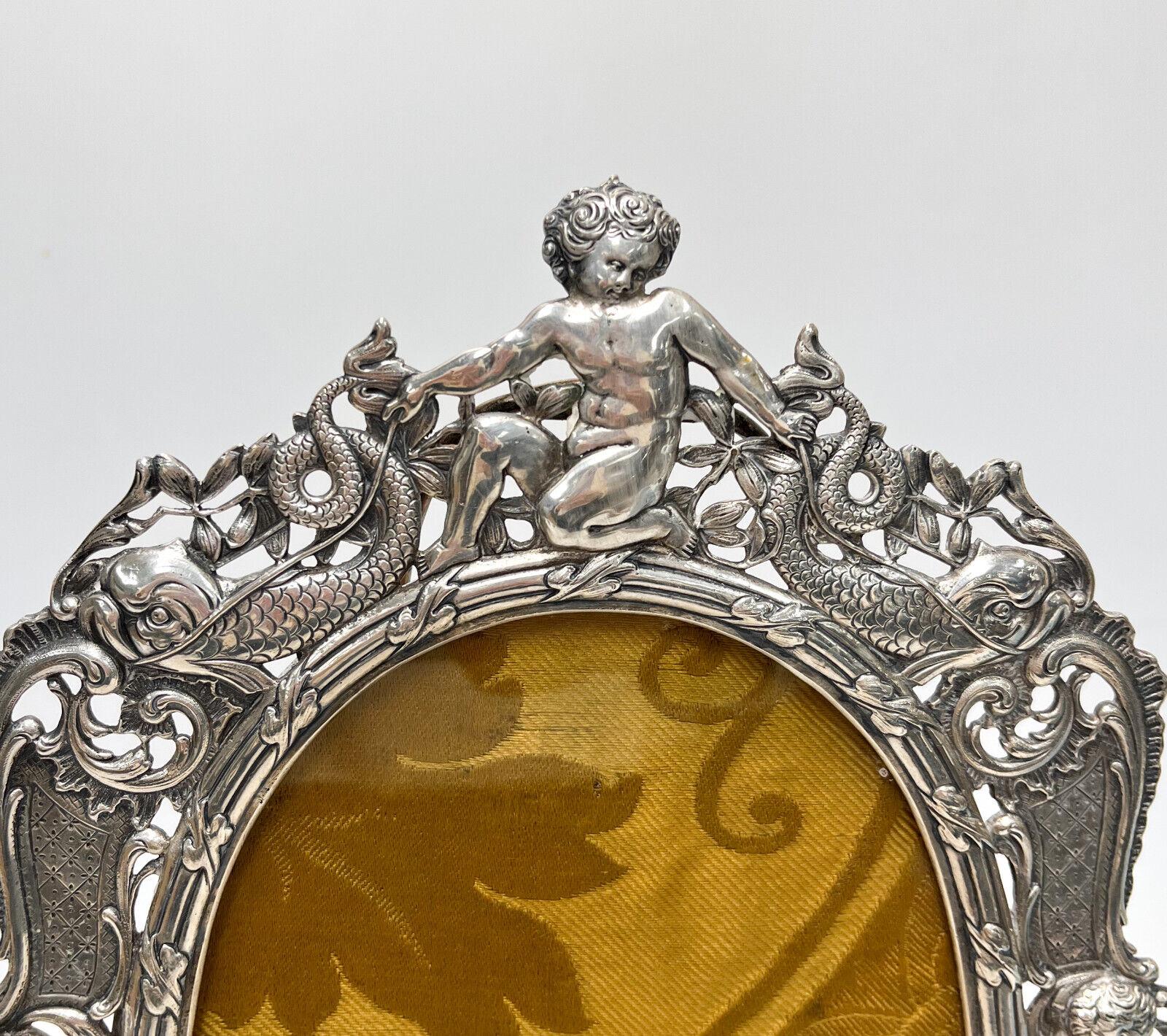 Continental 800 silver pierced cherub photo frame, circa 1900. Repousse figural cherubs with dolphin and foliate swag decoration. Unmarked, but tests for around 80% silver. Gold fabric backing. Likely Italian.

Additional Information: 
Weight