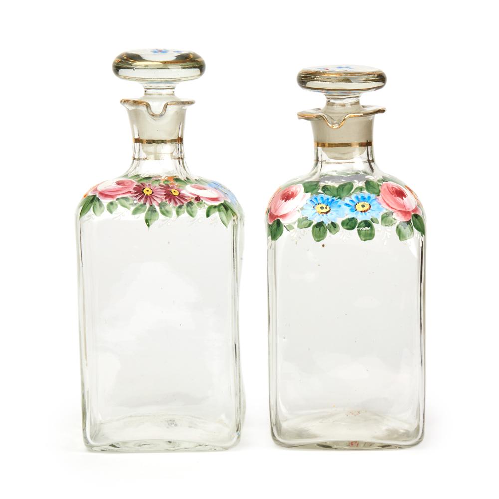 A fine pair, but individually made, antique continental clear glass decanters of square form hand painted with floral designs in colored enamels with narrow raised necks with pouring spout shaped rims and with hollow blown flat stoppers also