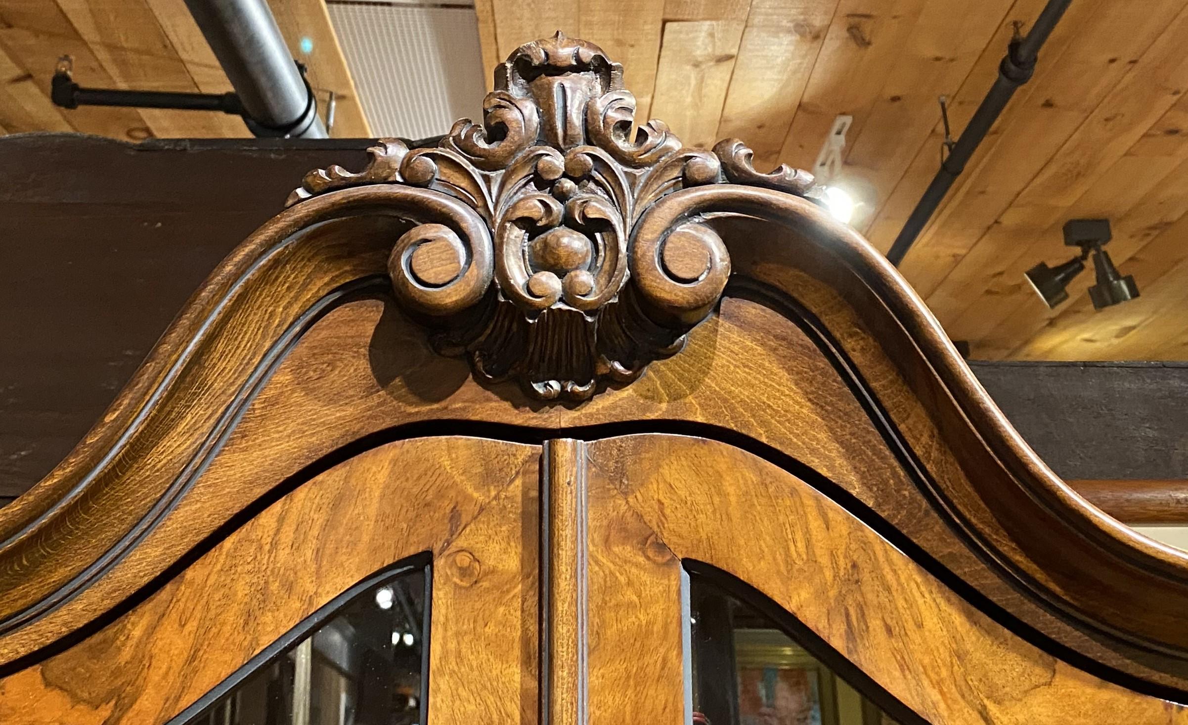 A fine example of a Continental Baroque Revival burled walnut one piece cabinet in a bombé form with arched carved foliate crest surmounting two conforming glass glazed doors which open to a two shelf interior, side glass panels, over three long