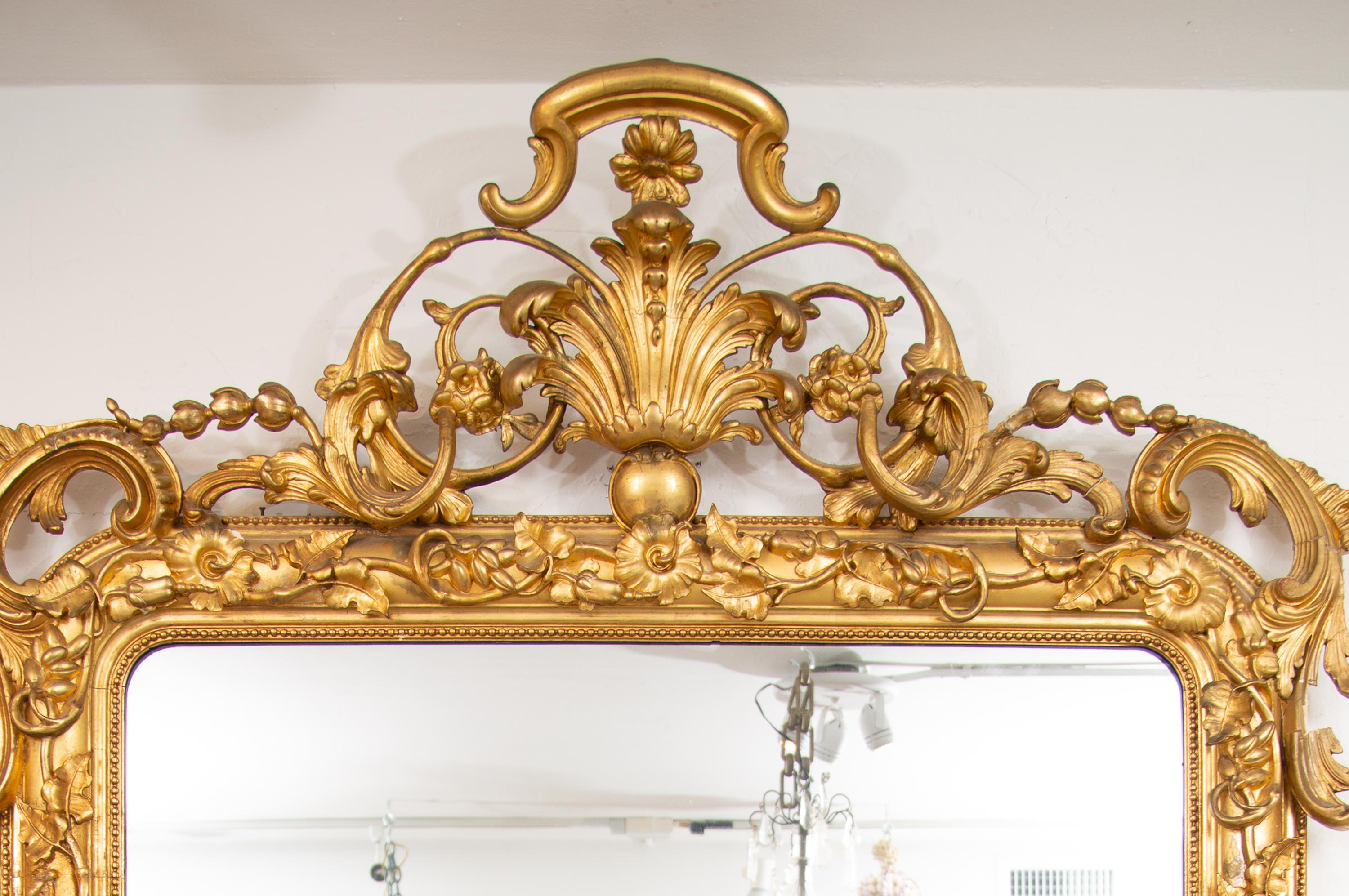 This is a large scale Continental carved giltwood mirror with a prominent arched decorative cornice over a frame intricately carved and gilded. There is an overall design of vines and flowers surrounding a large mirror plate, 19th century.