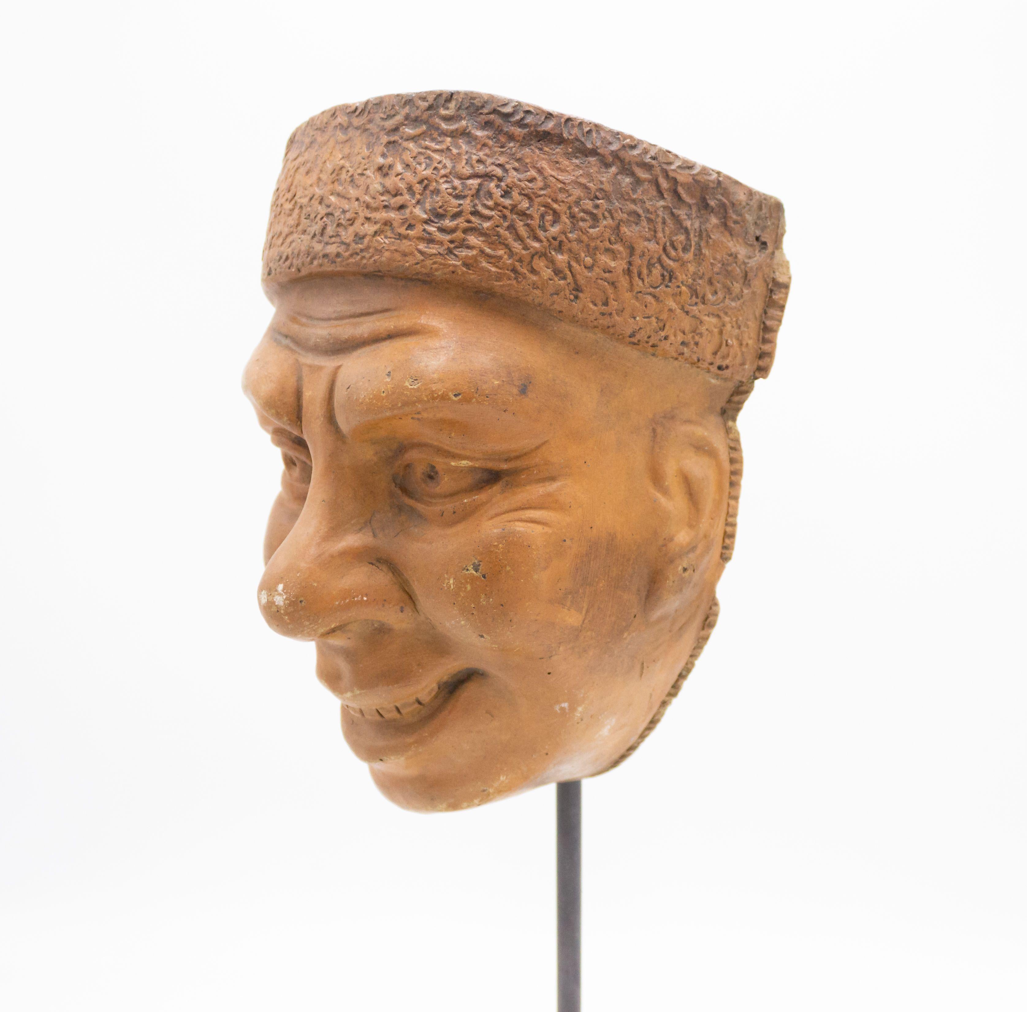 Continental German (late 19th Cent) sculpted terra-cotta master mask mold of a smiling face with a lamb's wool hat displayed on a square black marble base Stand (part of a collection).
  