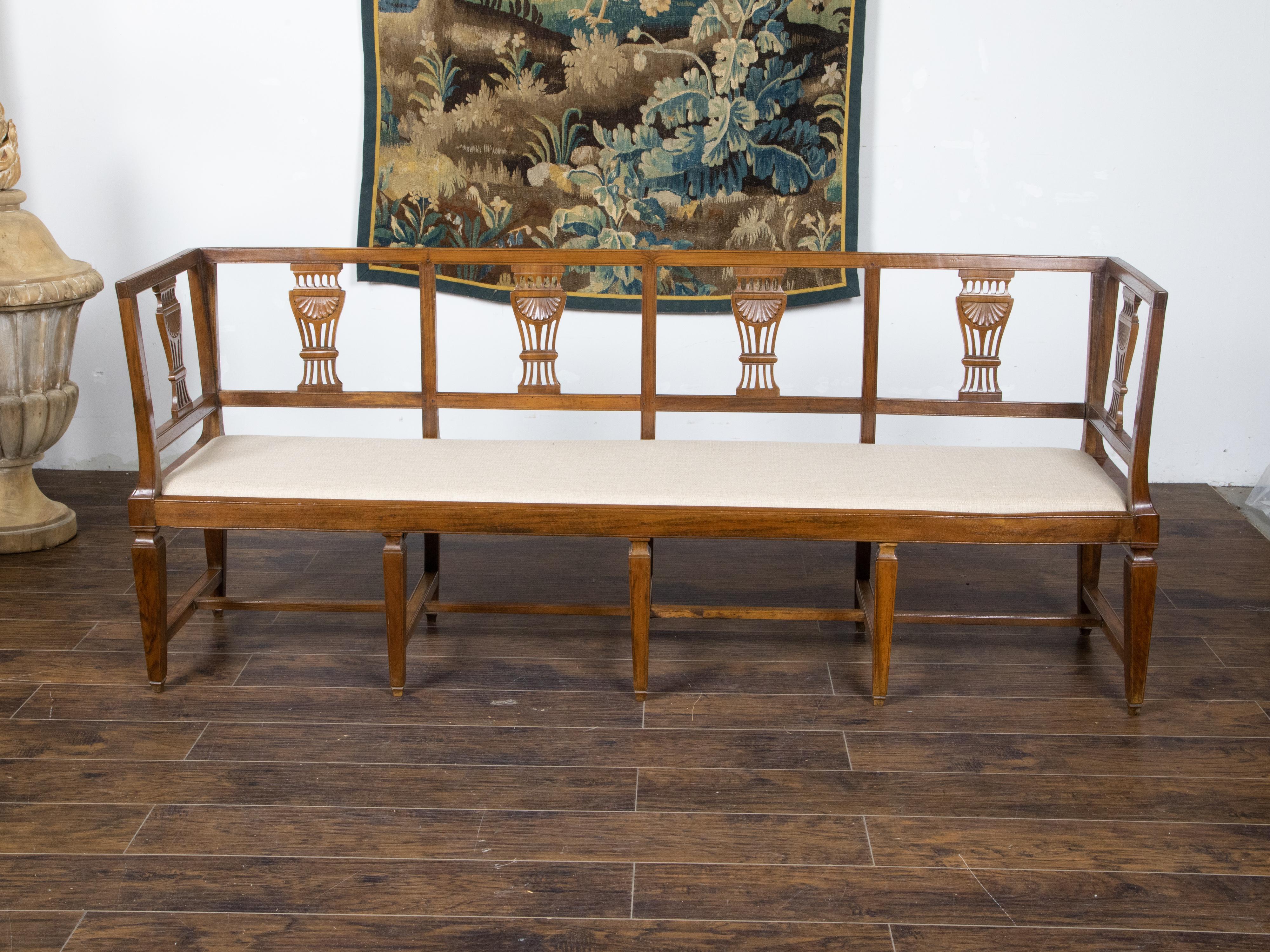 A continental Neoclassical style walnut four seater settee from the 19th century, with carved splats, upholstered seat, tapered legs and H-Form cross stretcher. Created in Continental Europe during the 19th century, this Neoclassical style walnut