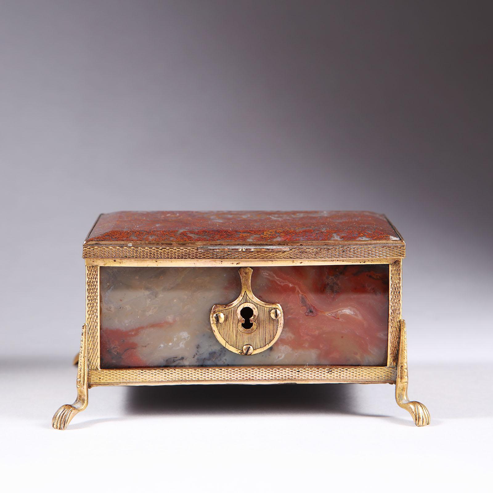 A fine 19th-century polished agate and gilt copper mounted box, all six sides with beveled panels of polished agate set within a tooled and gilt metal frame, the front detailed with a large tooled escutcheon. With a hinged lid and standing on curved