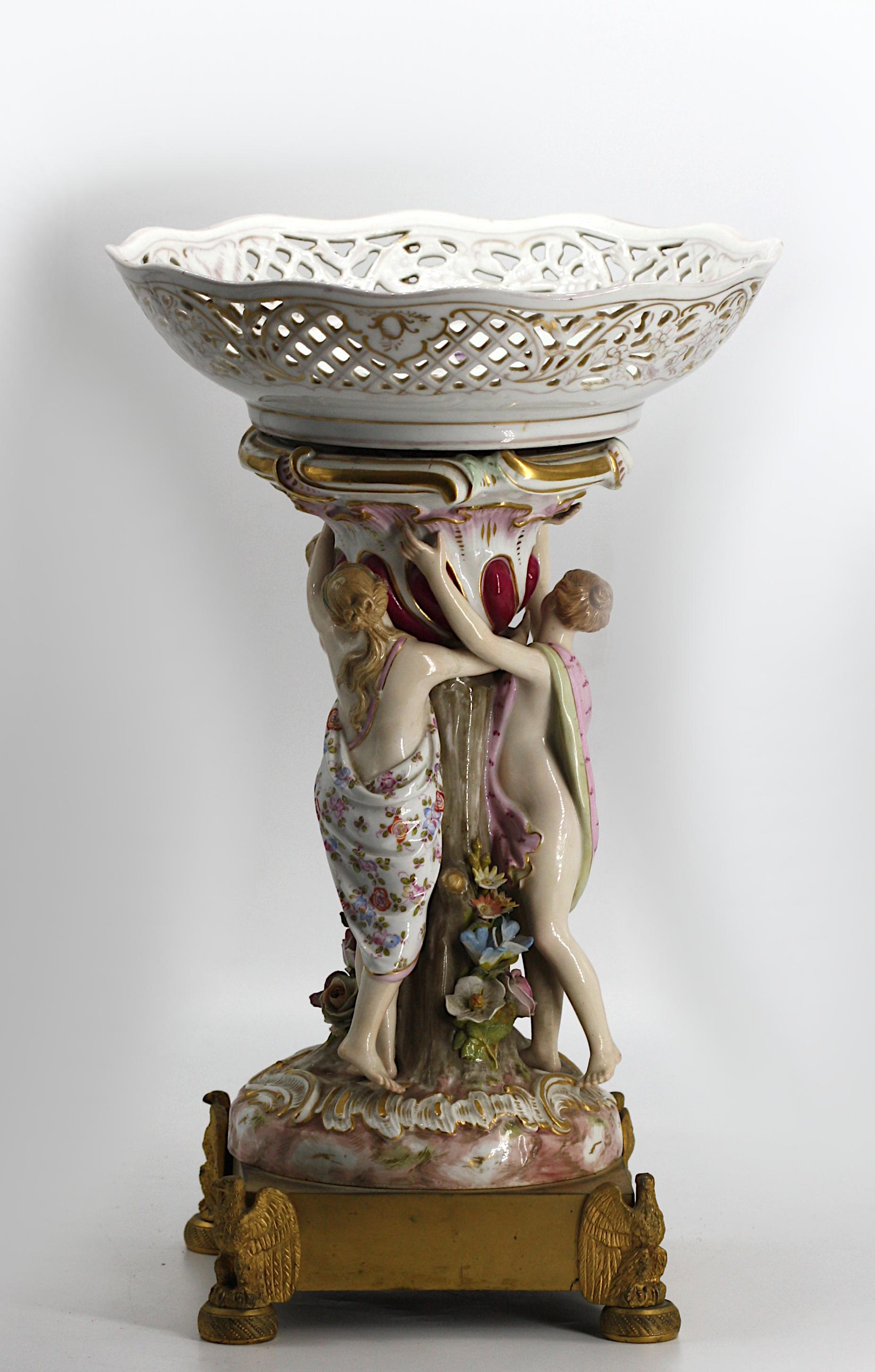 
Continental Porcelain Figural Centerpiece No visible marks. Circa 1900. Modeled with three classical maidens scantily clad some painted with foliage, on a scroll decorated circular base, atop a bronze plinth, with four eagle feet, surmounted by a