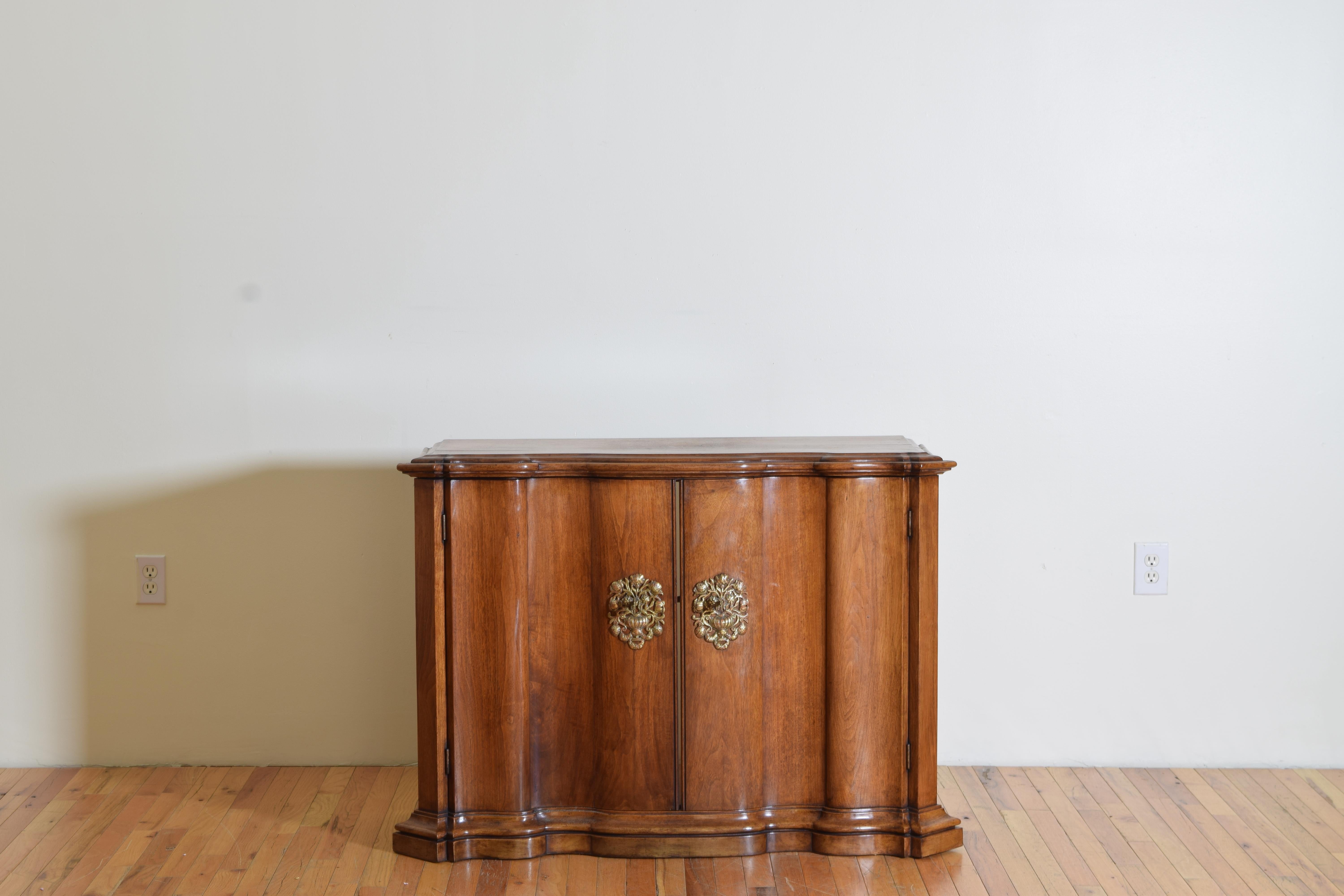 Having a shaped top with molded edge and serpentine shaped front atop a conforming case with two doors each having a large brass escutcheon plate, the interior with an open middle back flanked by two sets of shelves, raised on a graduated plinth