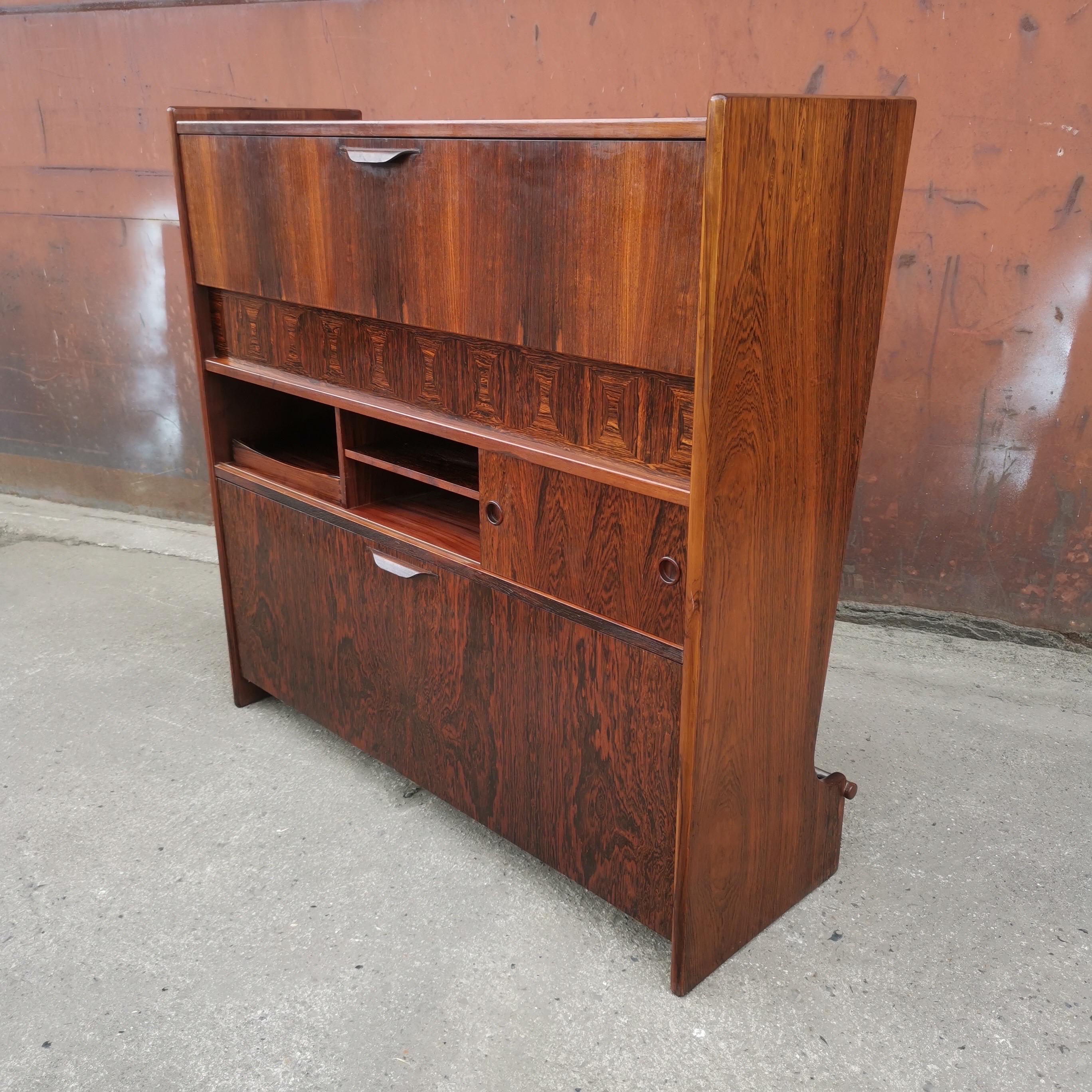 Danish designer Johannes Andersen created a functional yet decorative bar cabinet executed in rosewood. The closed front has a footrest whereas the functional side features various storage spaces. On top gives a downwards opening door access to a