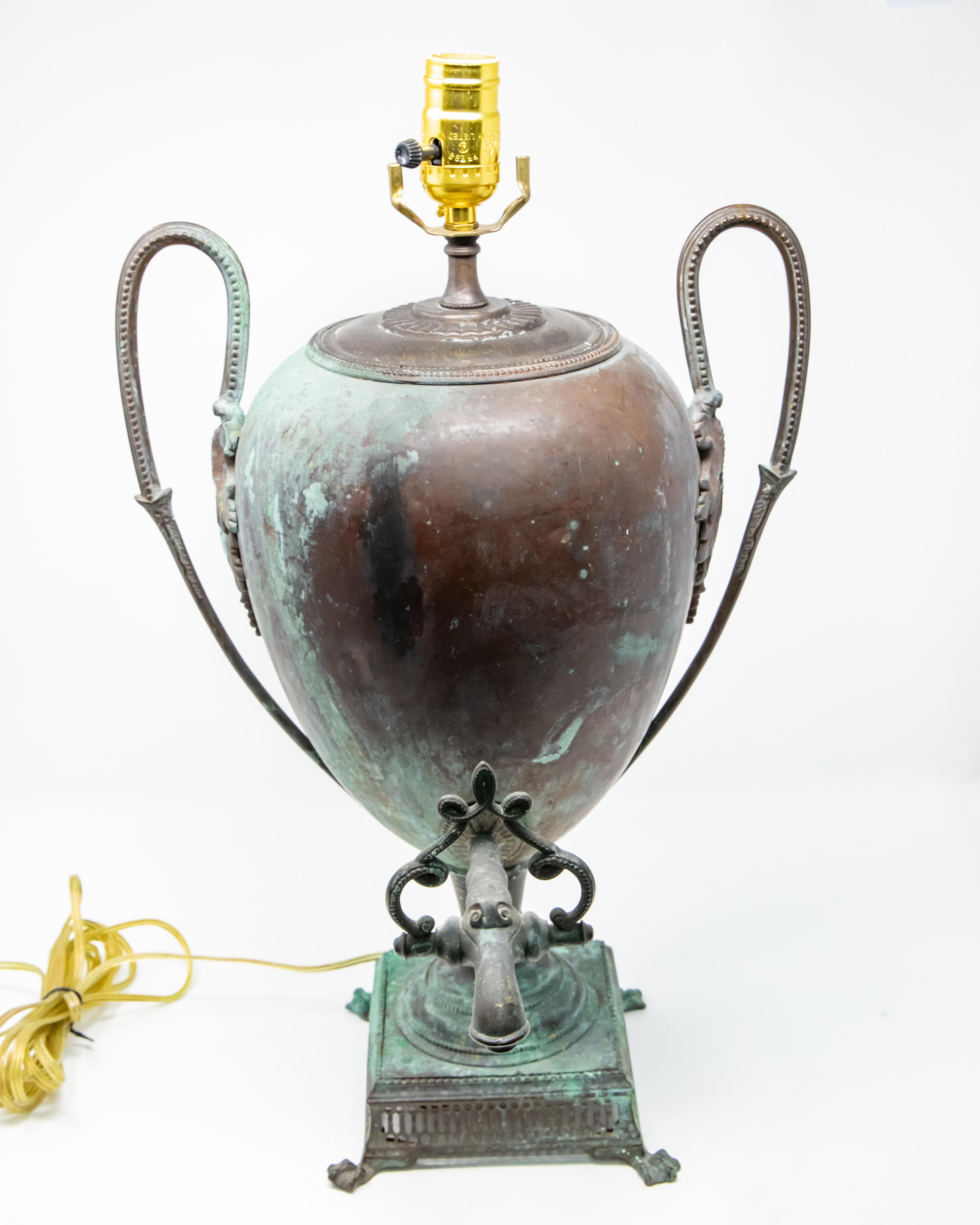 Offering this gorgeous patinated copper water vessel that was converted to lamp. The majestic shape of this old urn is breathtaking. With a tall looped handle with foliate detail that attaches to the side of urn. Sitting on a reticulated base on paw
