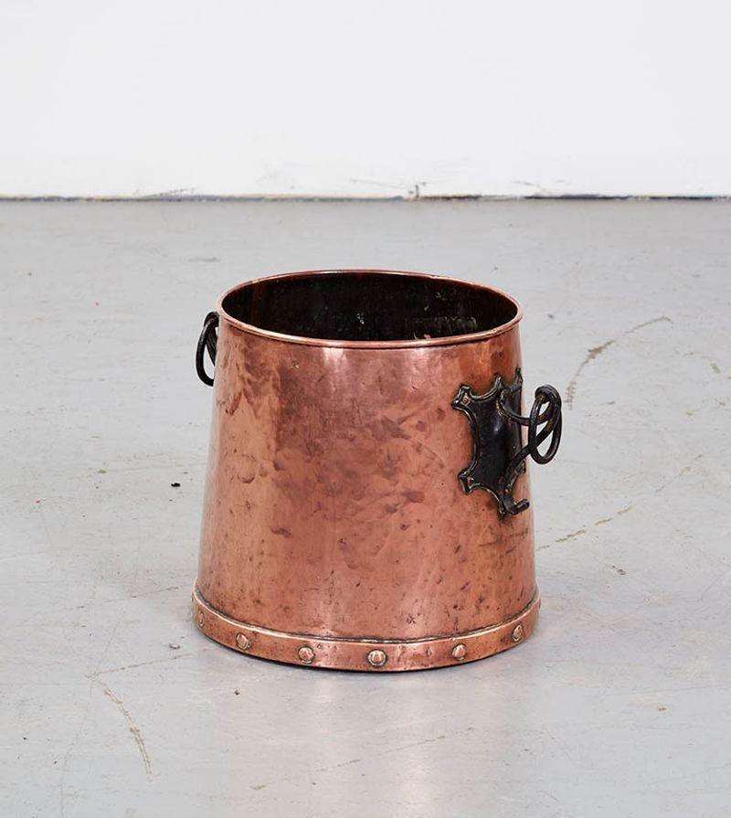 An Arts & Crafts copper bucket having rolled rim and slightly tapered profile with blackened wrought iron escutcheon and ring handles. Now useful for kindling next to a wood fireplace or as a decorative accessory next to a gas fireplace.