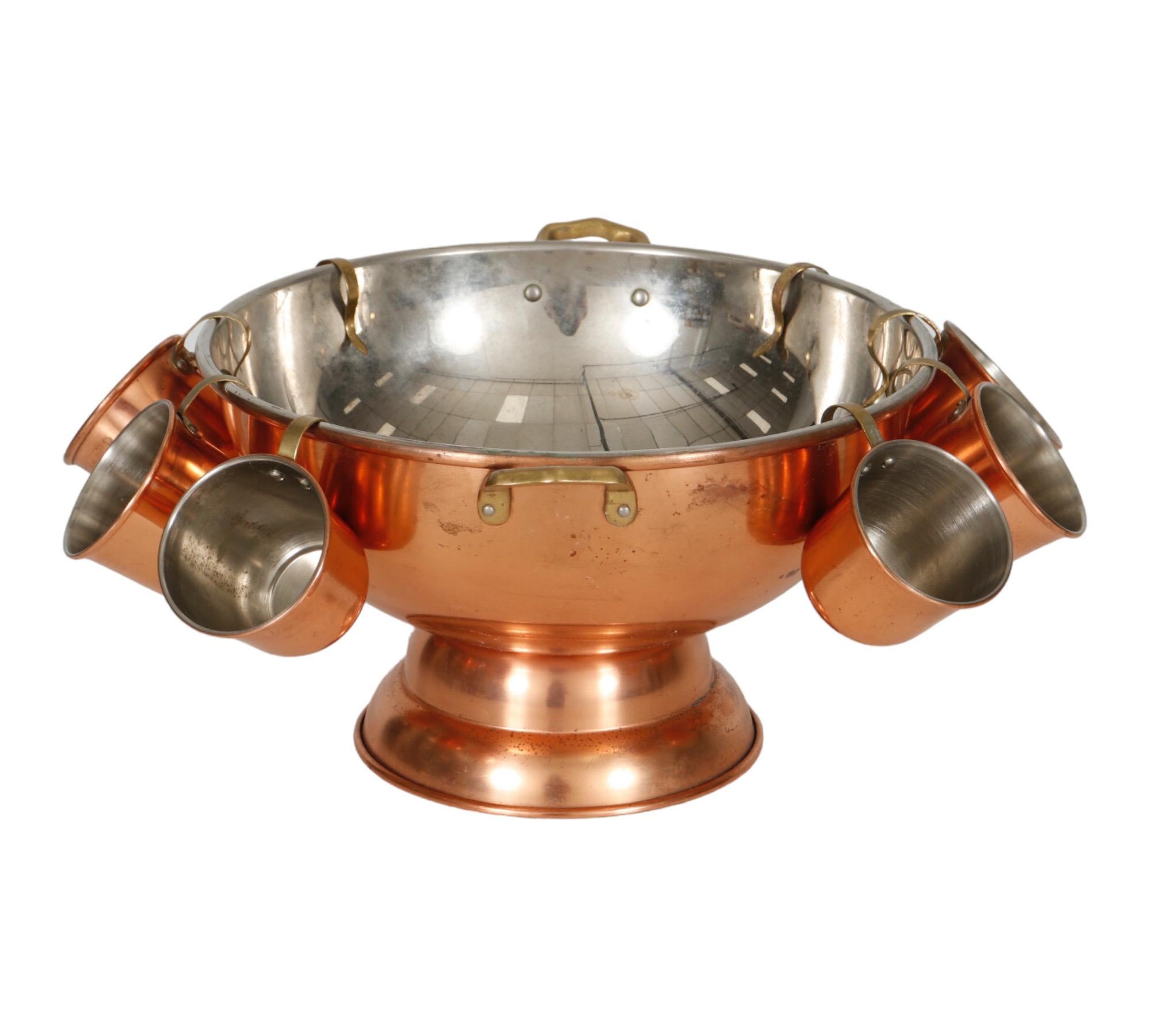 A copper punch bowl with a stainless-steel interior and two brass handles. Accompanied with eight copper mugs, also with stainless steel interiors, that hang on the edge of the bowl with brass handles. Bowl measures 12
