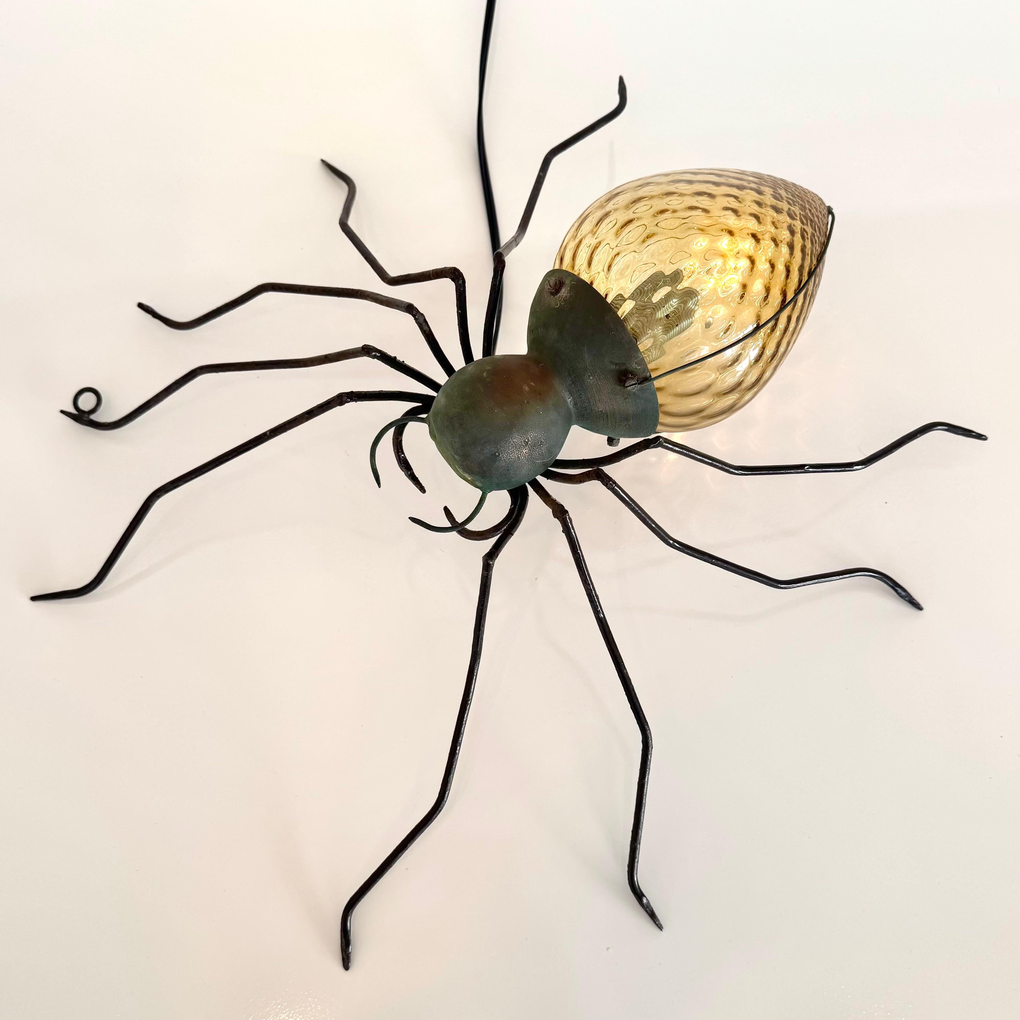 Unica applique a ragno in rame e metallo realizzata a mano in Italia. Grande presenza e dettaglio. Corpo in rame martellato con testa nera e gambe lunghe e sottili. L'addome è un bellissimo globo di vetro ambrato con una superficie piena di bolle.